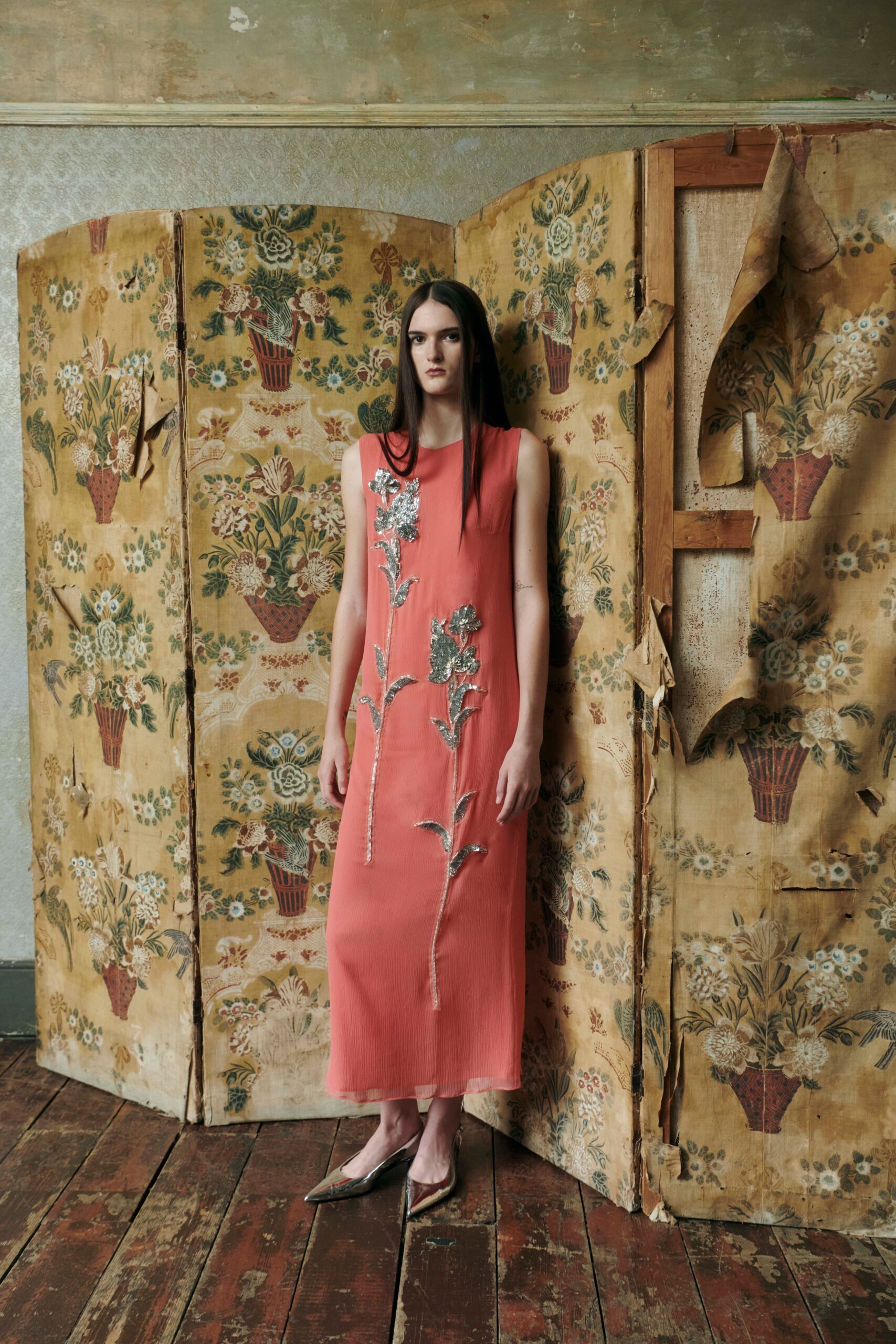 Coral sleeveless column dress with oversized silver floral paillettes climbing the front. Pointed mirror-leather slingbacks catching the floorboards in their toes. In a faded front room where the wallpaper has started peeling off the screen.