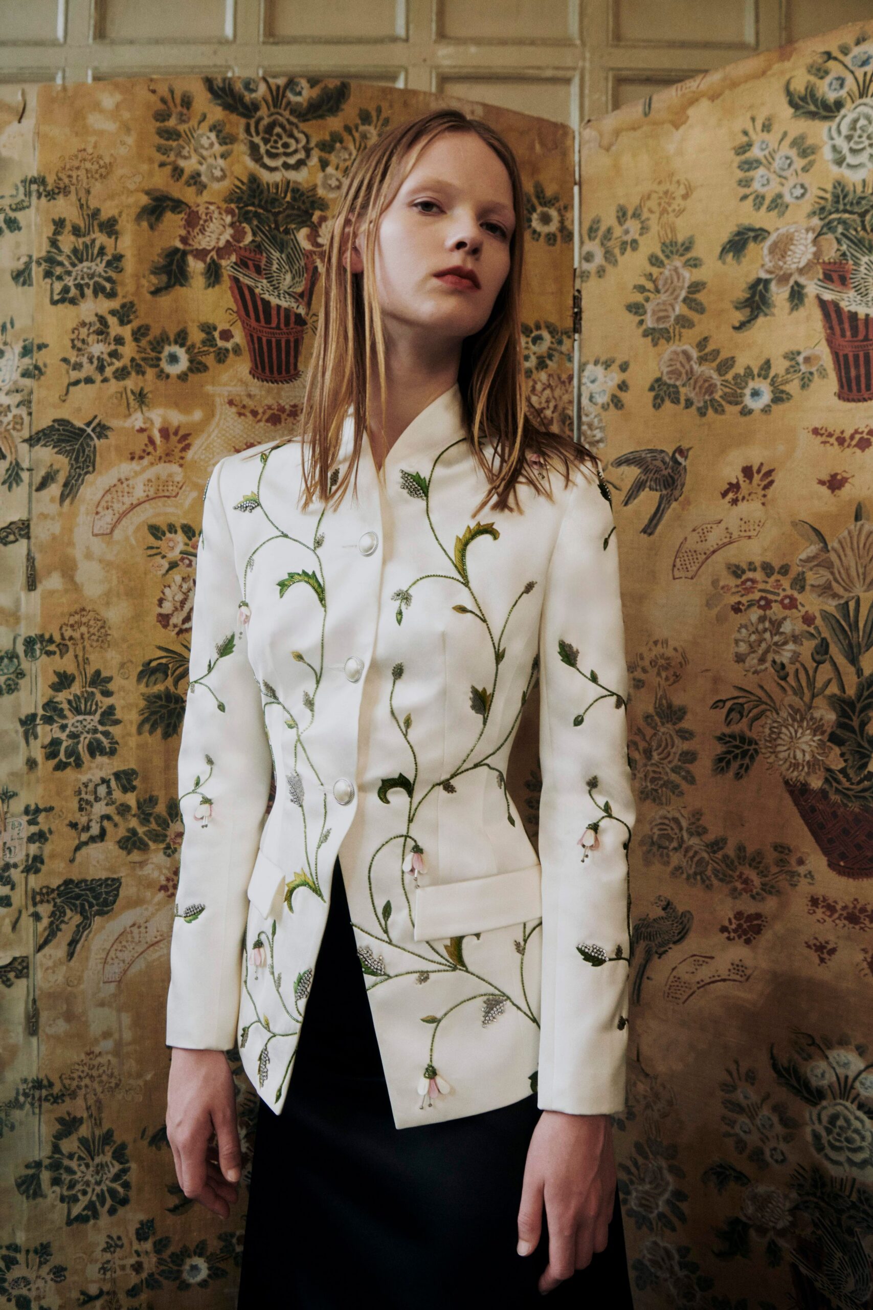 Ivory tailored jacket traced with green vine embroidery and pink bell flowers. Black trousers disappearing under the long peplum-like front panel. In a drafty drawing room while the radiator knocks before dinner.