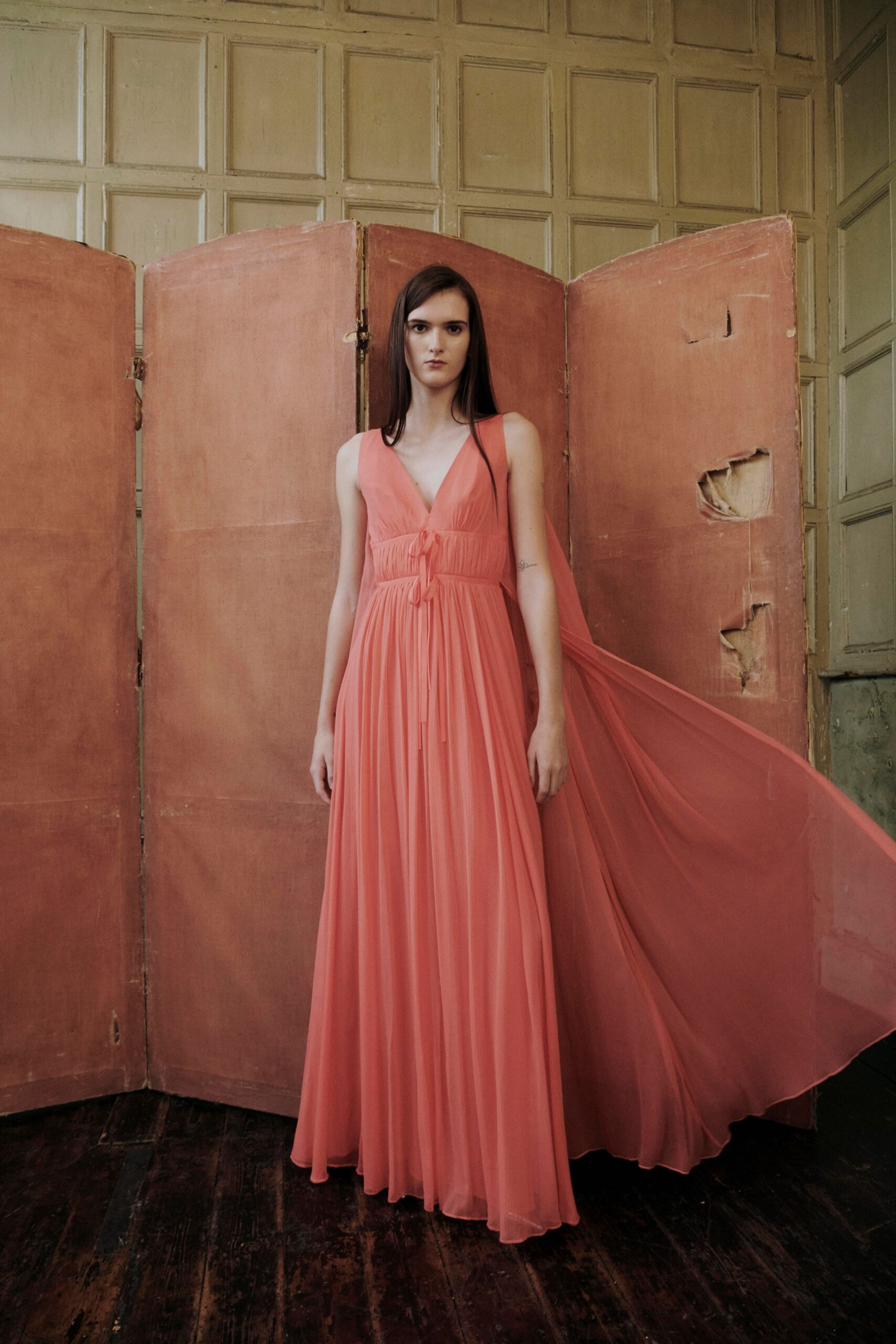 Coral chiffon gown with knife-pleated skirt and long back panel drifting sideways. Caught in a drafty manor hallway while someone opens the wrong door.