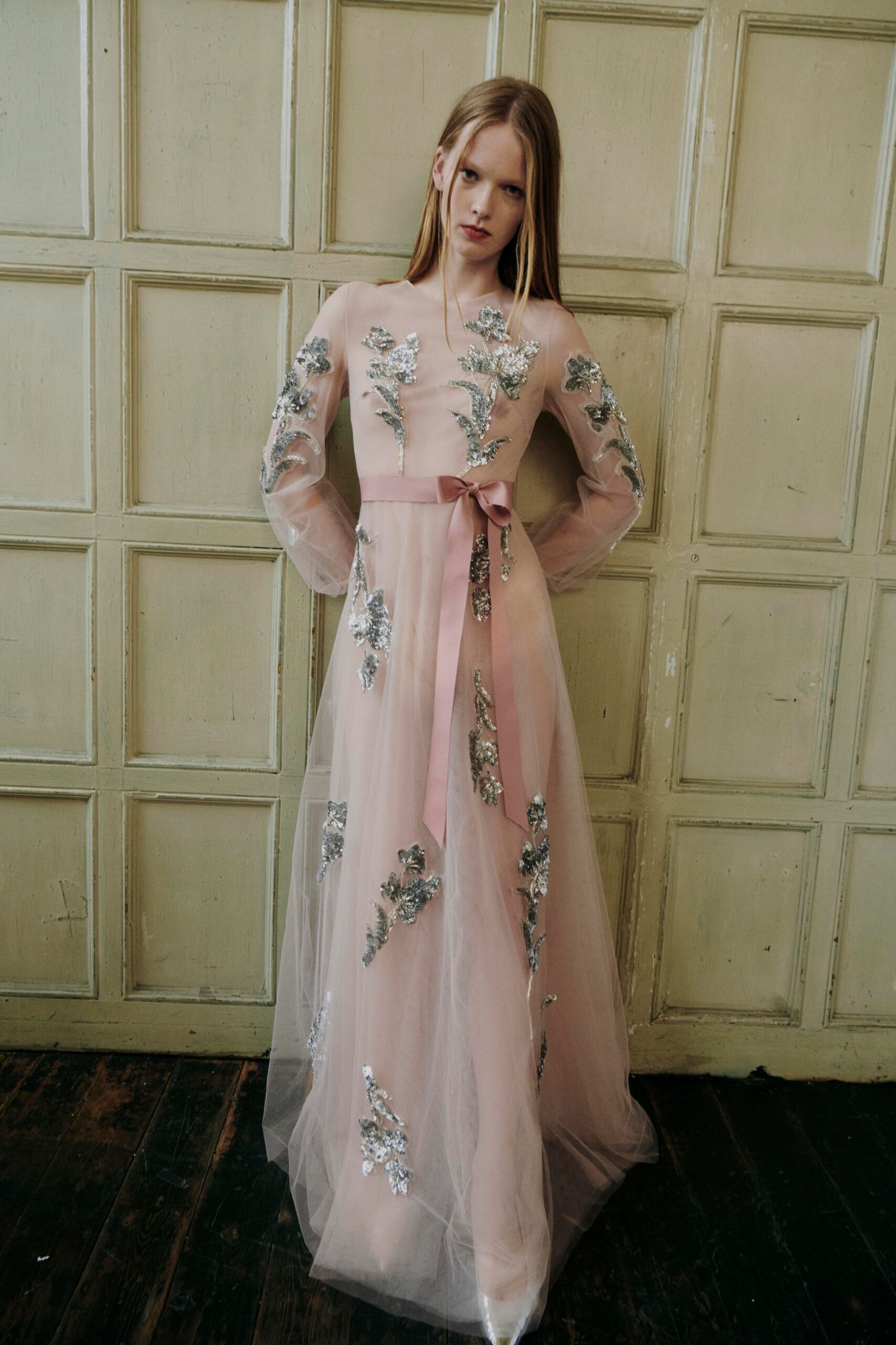 Blush tulle gown with silver floral appliques and a long satin waist bow. White pointed shoes dimly visible through the floor-length sheer hem. In a faded manor hallway before everyone is called down to dinner.