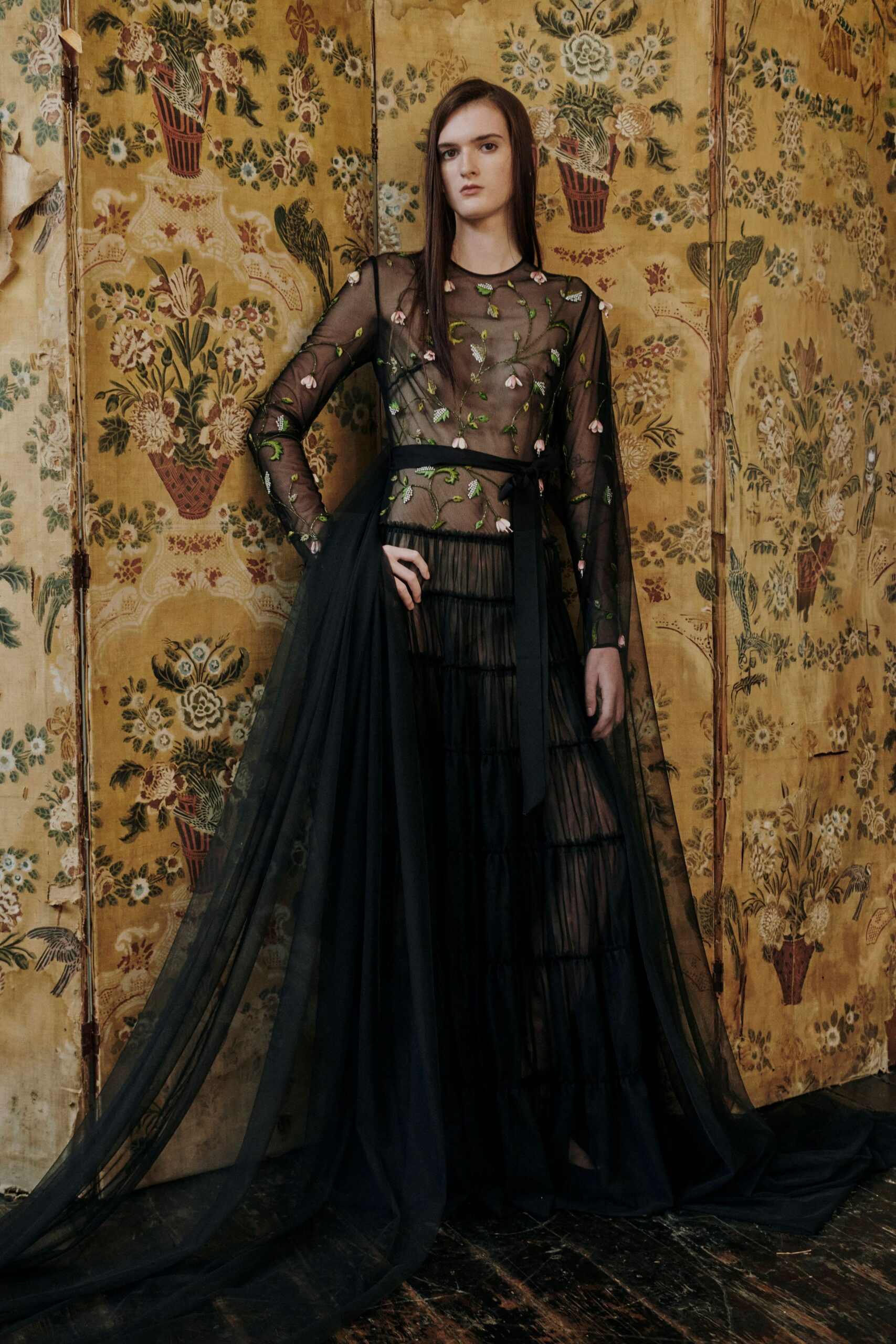 Sheer black tulle top embroidered with trailing green stems and pink buds. Black grosgrain sash tied low, one long end falling past the hip. Tiered black tulle skirt with narrow gathered bands showing through. In a faded music room after midnight, waiting for the windows to fog.