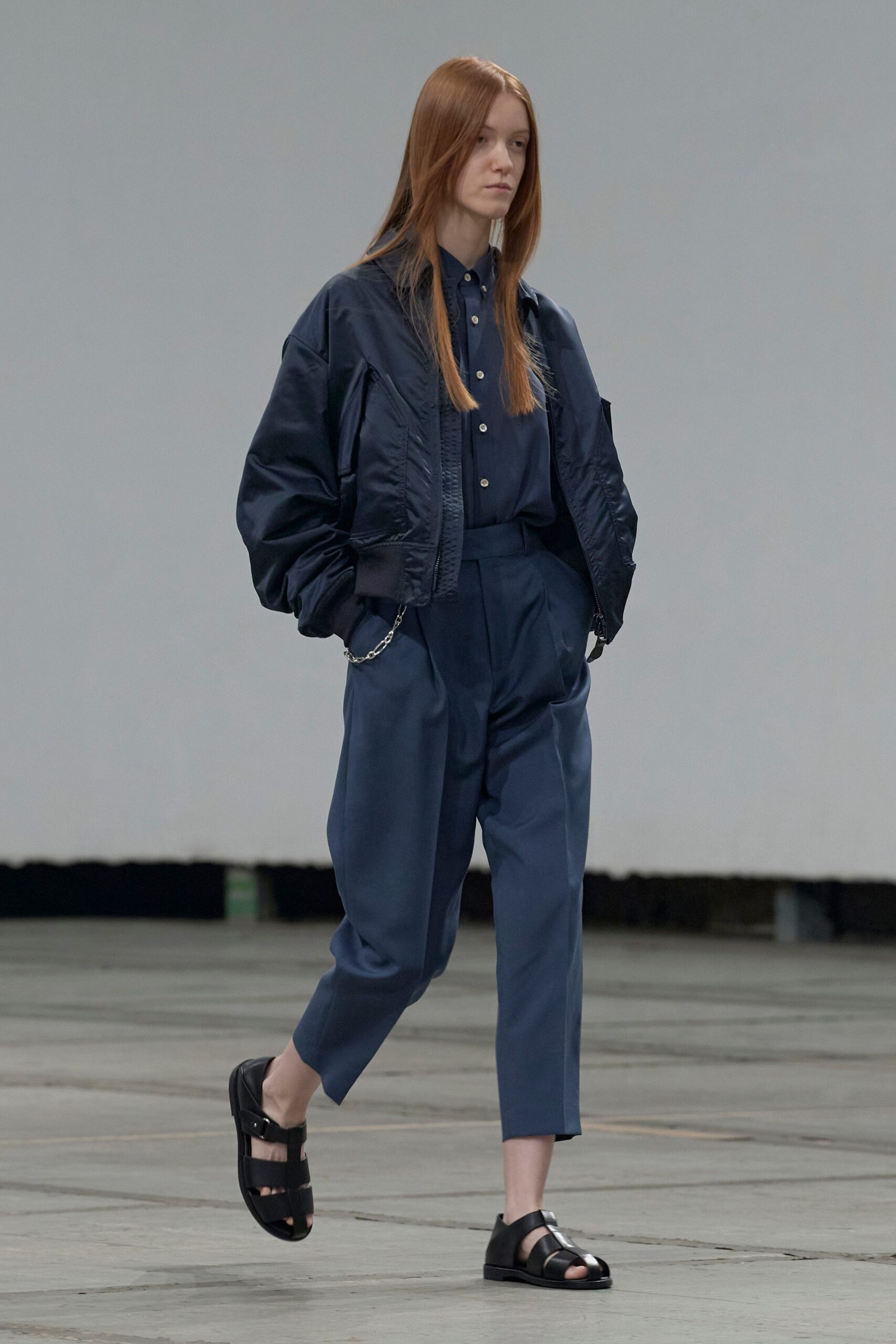 Ink satin bomber jacket, dropped shoulders and a chunky black zip. Matching navy button-down, top button fastened under a neat collar. High-waisted slate trousers with deep front pleats and a silver hip chain. Black cutout leather sandals with a closed heel. Crossing an empty ferry terminal before the first announcement.