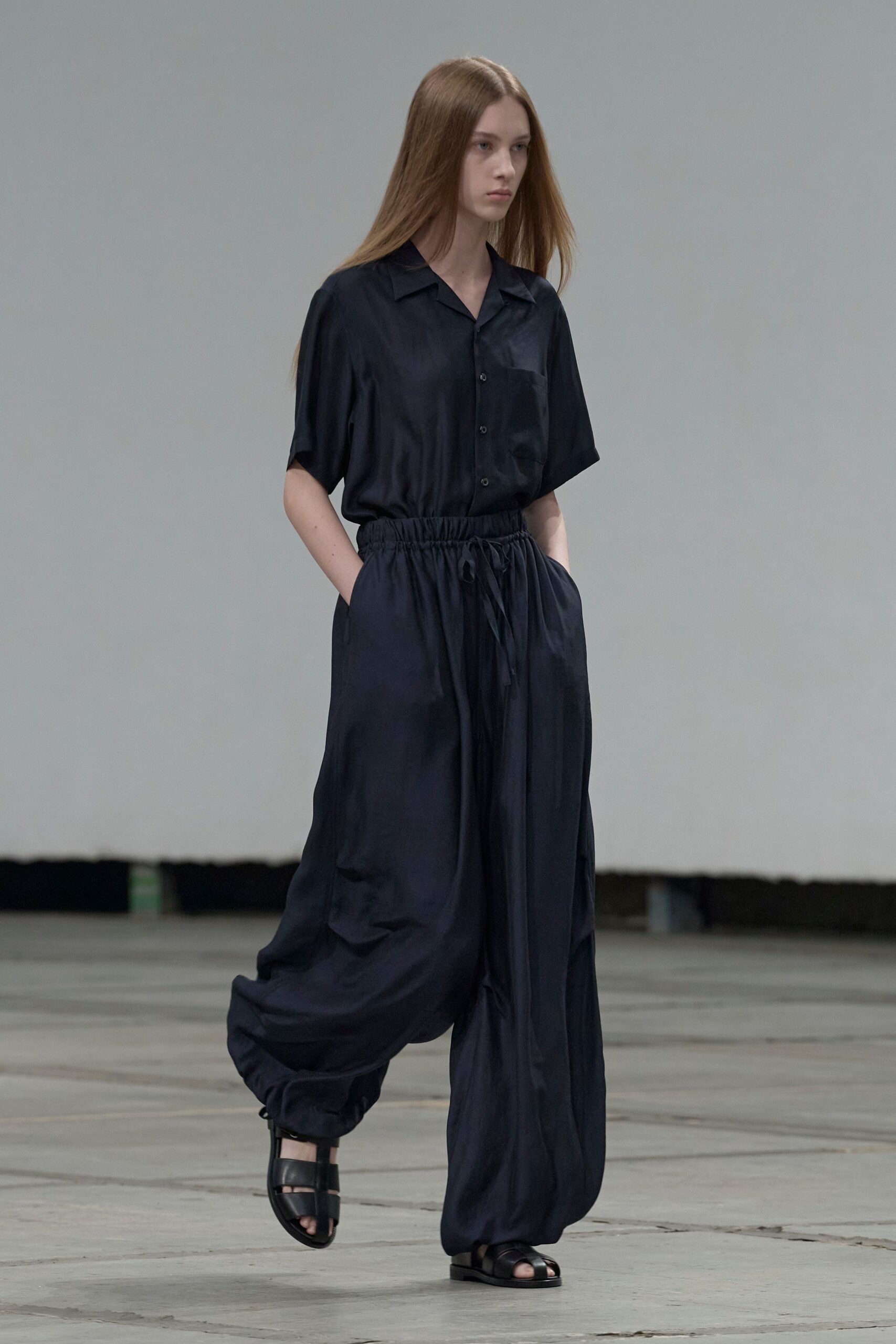 Ink-dark satin camp shirt, one chest pocket and small black buttons. Matching satin trousers, elastic drawstring waist and softly ballooned cuffs. Black leather fisherman sandals. For crossing a silent terminal before the first departures board flickers on.