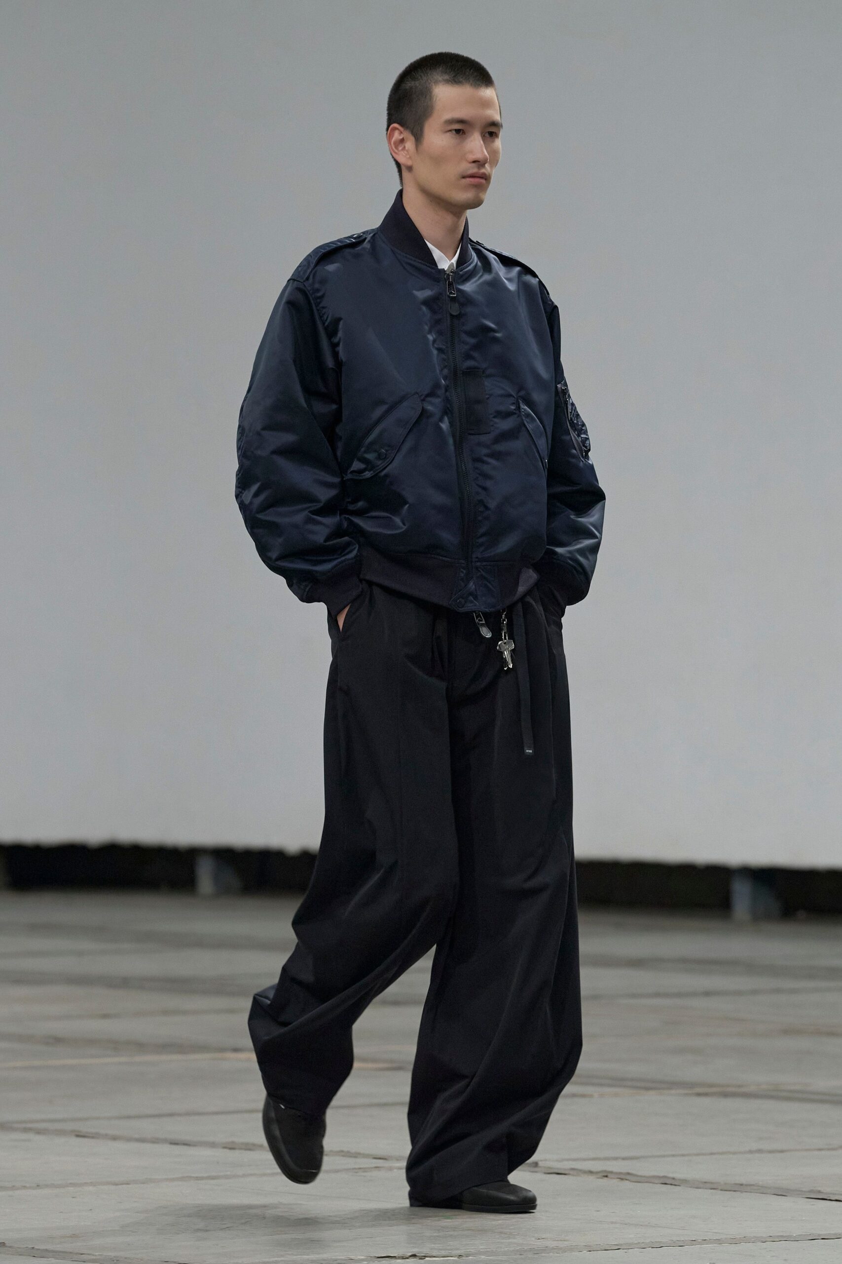 Ink navy satin bomber with epaulettes and a ruched sleeve pocket. White pointed collar peeking cleanly at the throat. Black trousers cut full and long, keys swinging from the drawstring. Square-toe black leather shoes. Crossing an empty aircraft hangar before the rain starts.
