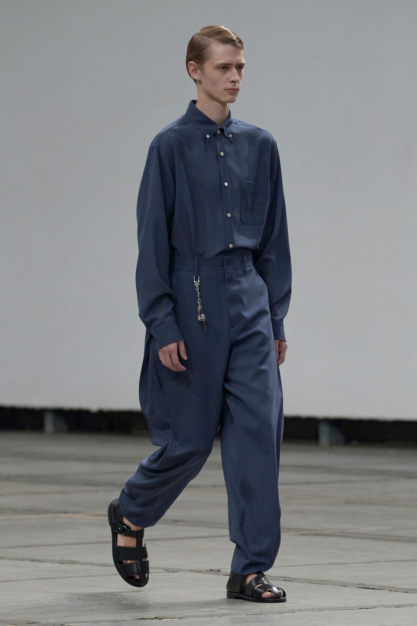 Slate-blue button-down in fluid cloth, chest pocket set slightly low. Matching high-waisted trousers cut full through the hip, tapering softly. Silver clasp with two keys swinging from the waistband. Black leather fisherman sandals with broad bands over the toes. Crossing an empty civic hall just after the cleaners finished.