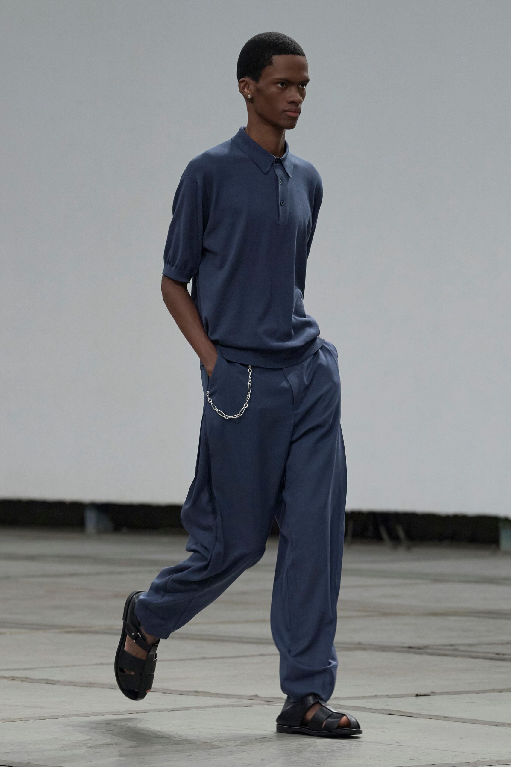 Slate-blue fine knit polo, three dark buttons under a soft collar. Matching draped trousers with a silver wallet chain at the hip. Black leather fisherman sandals with broad cutout straps. Crossing an empty convention hall before the doors open.