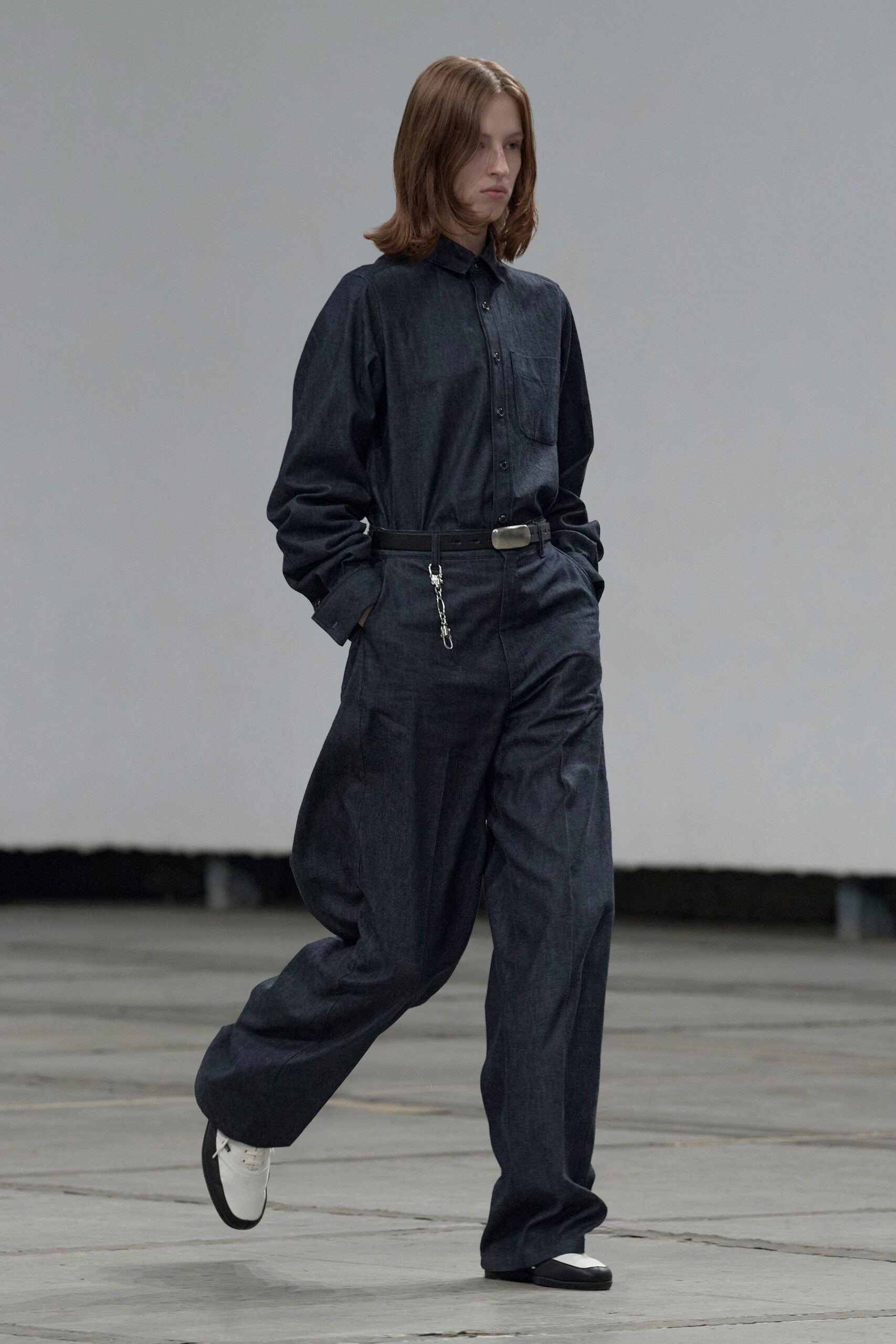 Dark indigo chambray shirt with a single rounded chest pocket. Black leather belt with a blunt silver plaque buckle and chain clip. Matching chambray trousers cut full through the leg, breaking over the shoe. White leather lace-ups with black rubber toe caps. Crossing an empty depot floor before the first delivery trucks arrive.