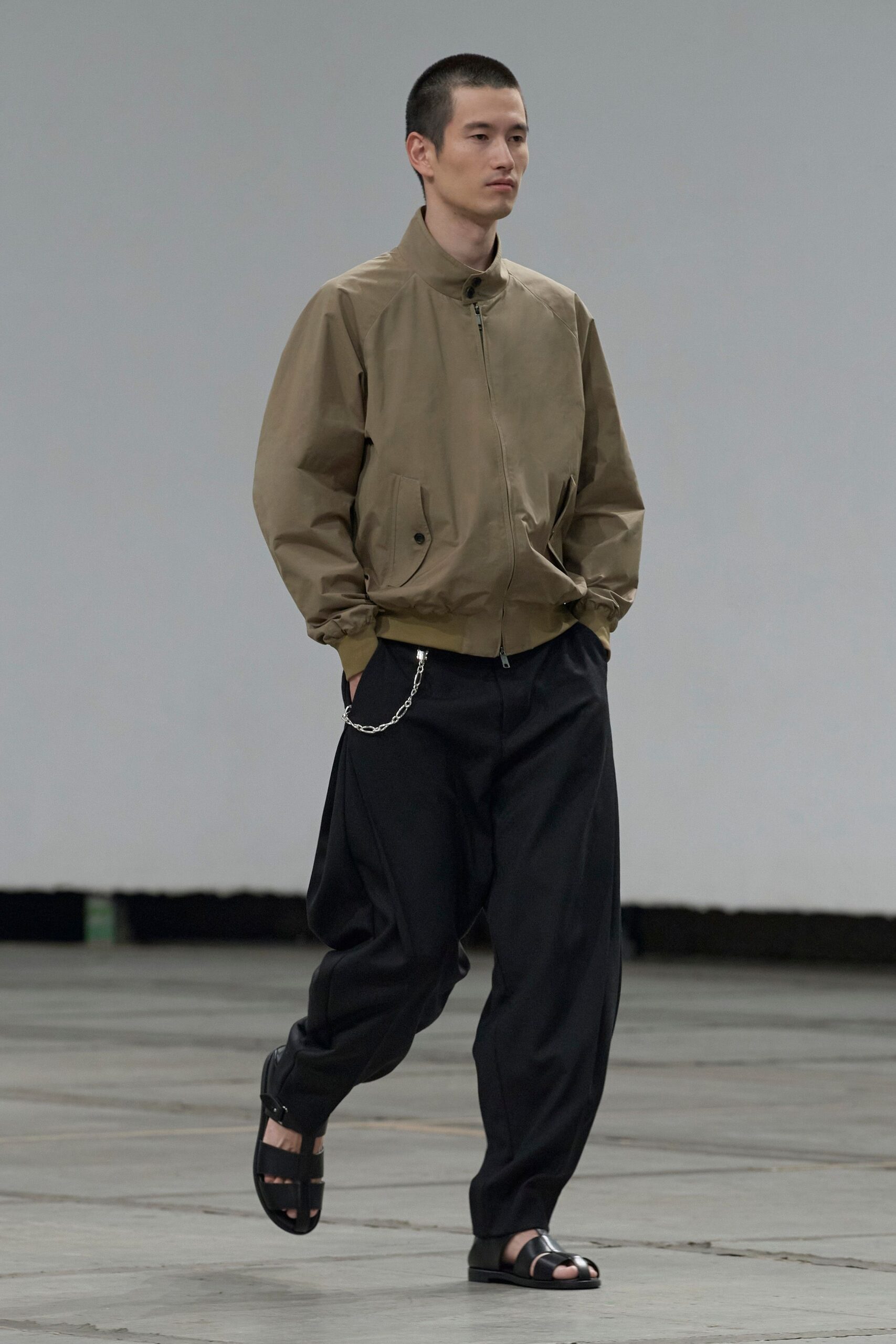 Taupe zip-front blouson with a stand collar and buttoned flap pockets. Black pleated trousers cut loose through the leg, silver wallet chain at the hip. Black leather fisherman sandals with a broad toe strap. Walking across an empty terminal before the first departures board flickers on.