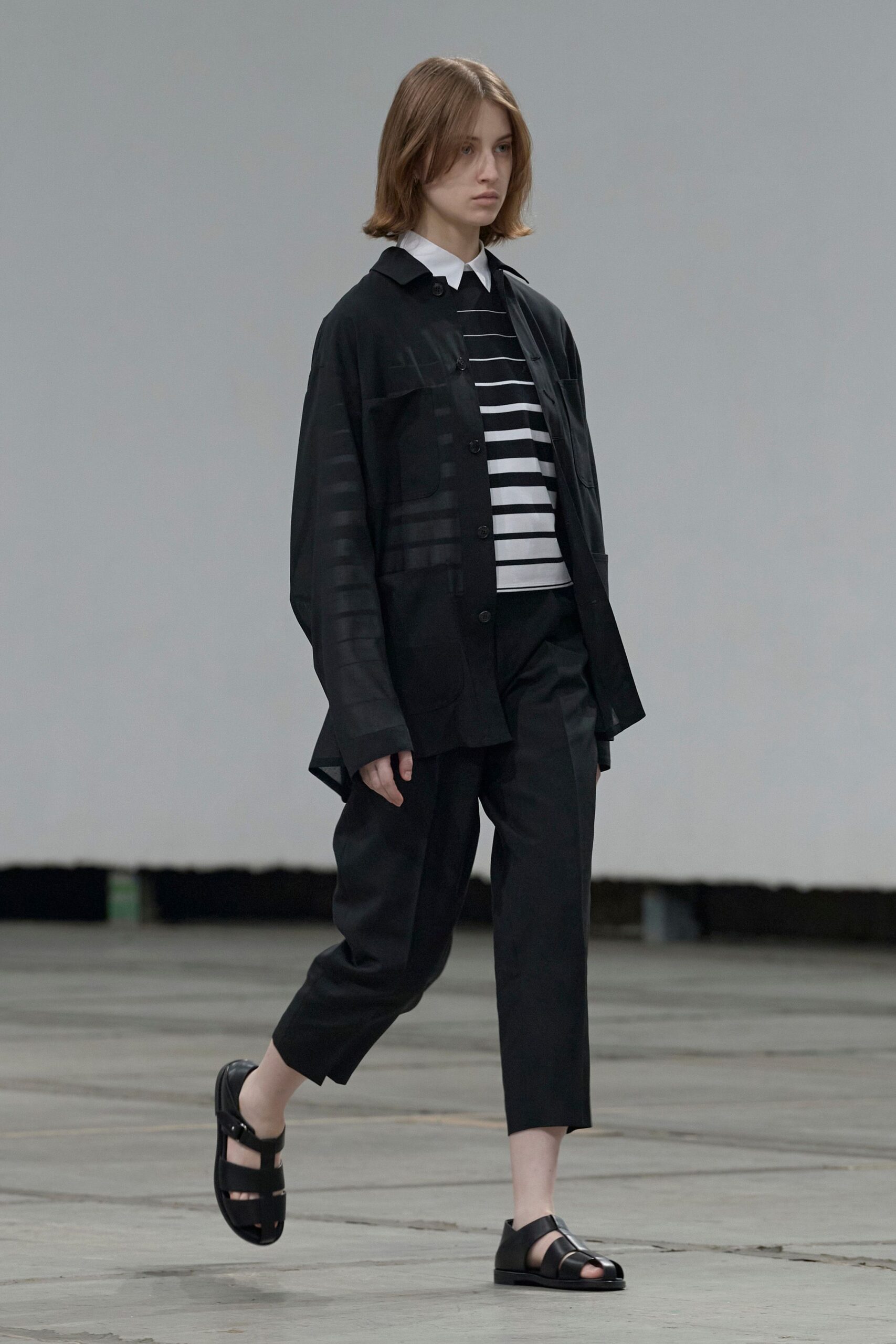 Sheer black coat with patch pockets and a dipped back hem. Black knit polo with white rugby stripes and a sharp white collar. Black cropped trousers, loose through the thigh and clean at the ankle. Black leather fisher sandals with broad caged straps. Crossing an echoing civic hall before the chairs are set out.
