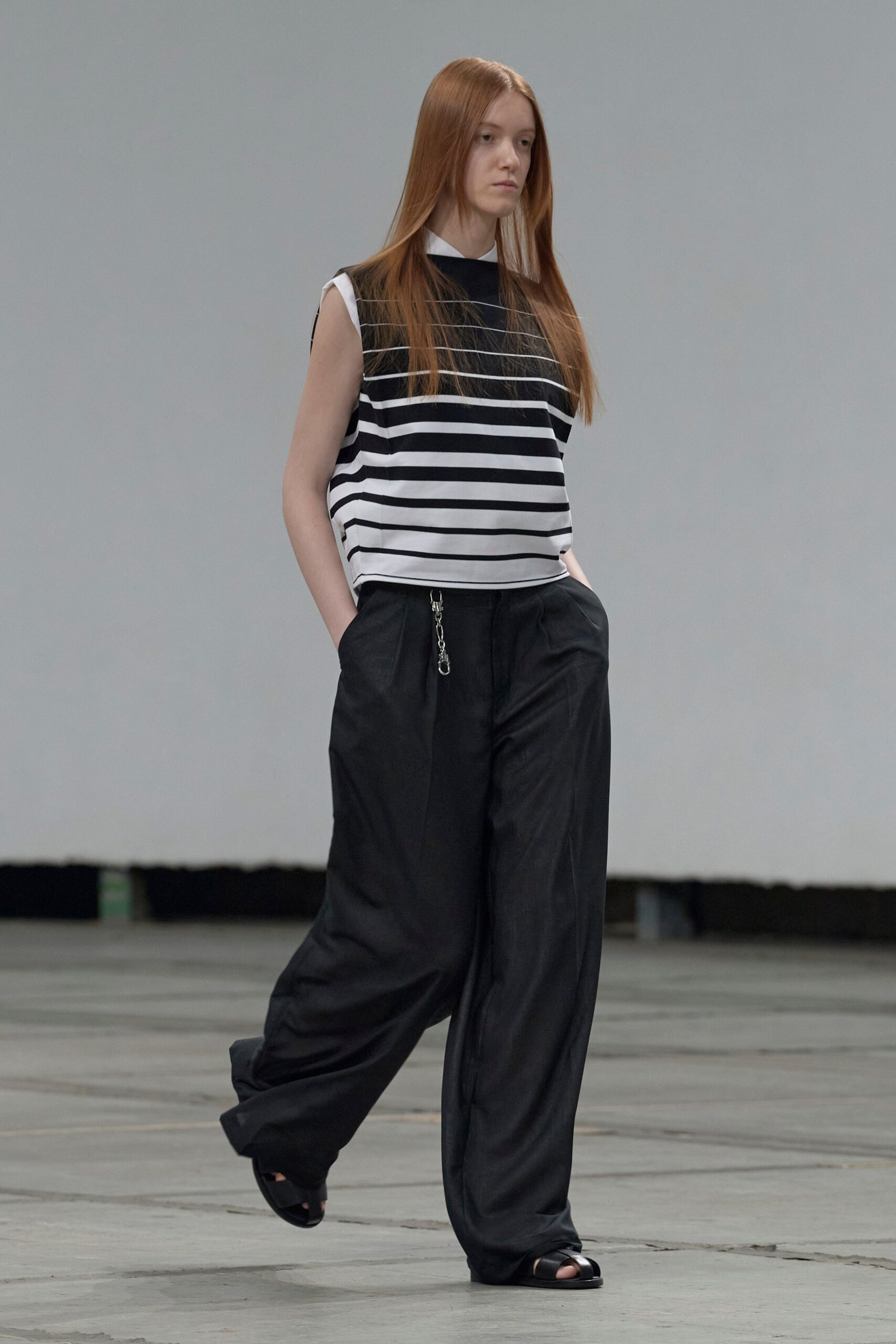 Boxy black-and-white striped shell, crisp white collar peeking at the neck. Black wide-leg trousers in fluid twill, silver clip hanging from the fly. Black leather slide sandals with a broad crossover strap. Walking the echoing concourse before the first train boards.