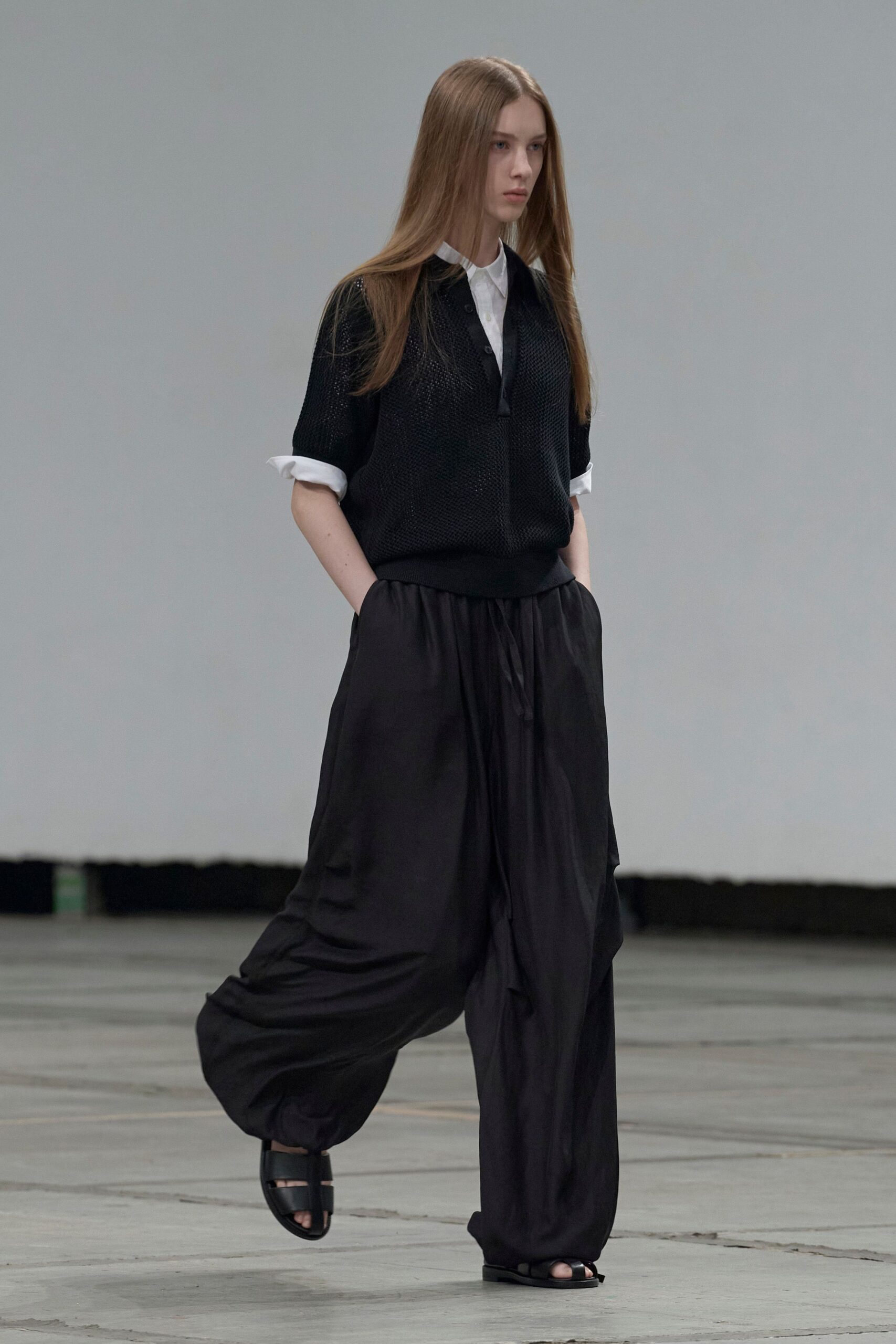 White poplin shirt with a small pointed collar and rolled cuffs. Black open-knit short-sleeve layer, placket hanging longer at the chest. Inky parachute trousers, gathered at the ankle with a loose drawstring. Black leather slide sandals with two broad straps. Walking across an empty hangar floor before the heat lifts.