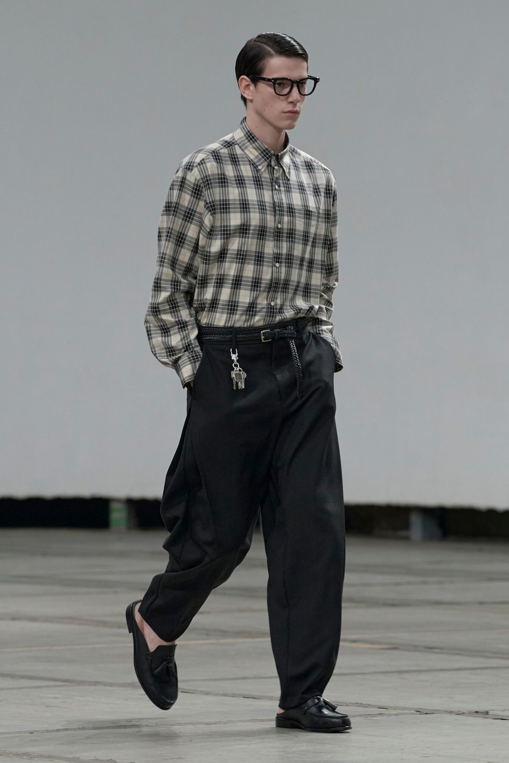 Thick black acetate frames. Grey and cream plaid cotton shirt, button-down collar pinned neatly. Black braided leather belt with keys clipped at the hip. Inky pleated trousers falling loose from a high waist. Black leather tassel loafers worn without socks. Walking into the office tower on Sunday to water someone else's plants.