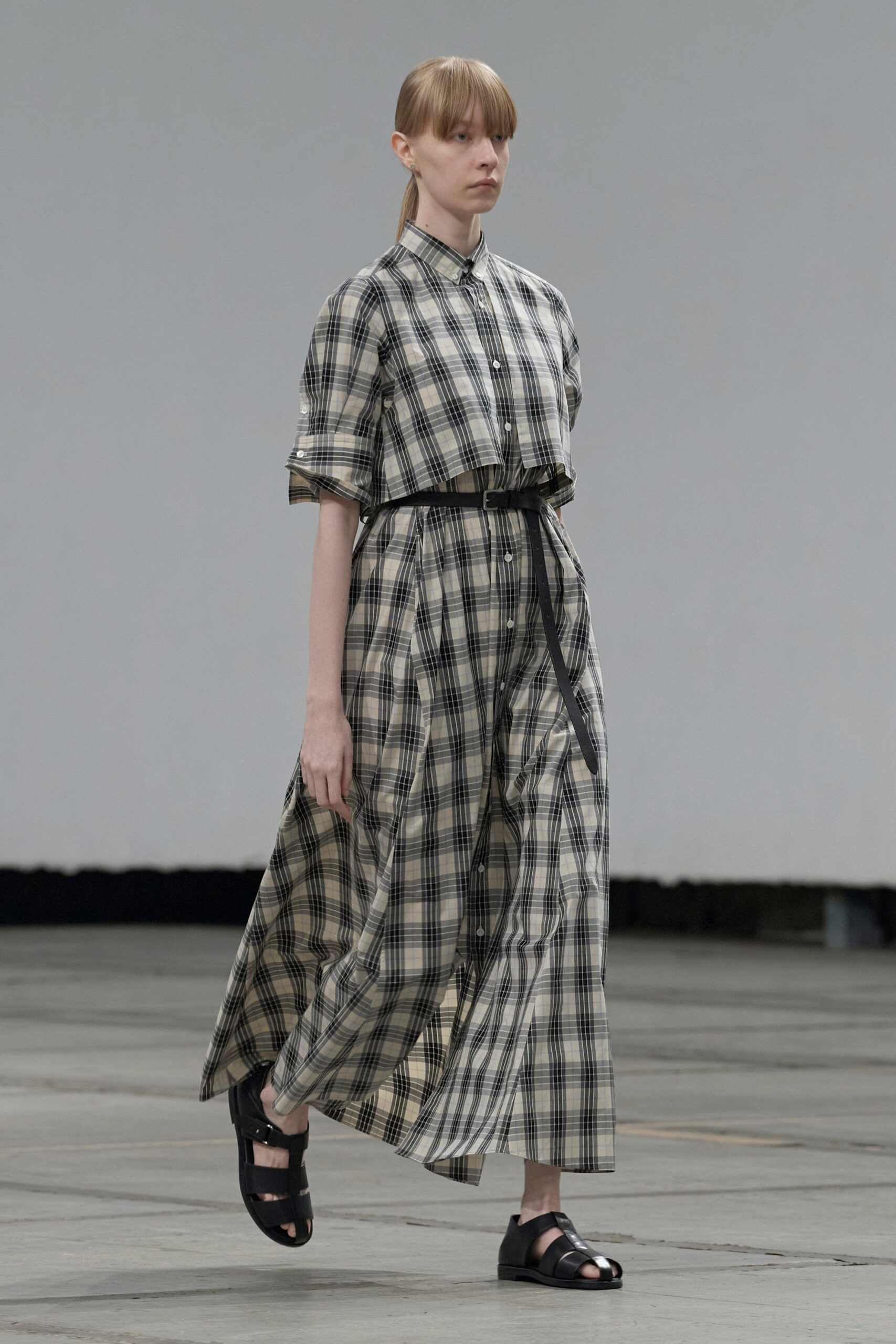 Stone and black plaid shirtdress with a cropped overlay panel at the bodice. Narrow black belt, long tongue hanging past the buckle. Black cutout leather sandals with broad straps over the toes. Walking across an empty civic hall after the folding chairs are stacked.