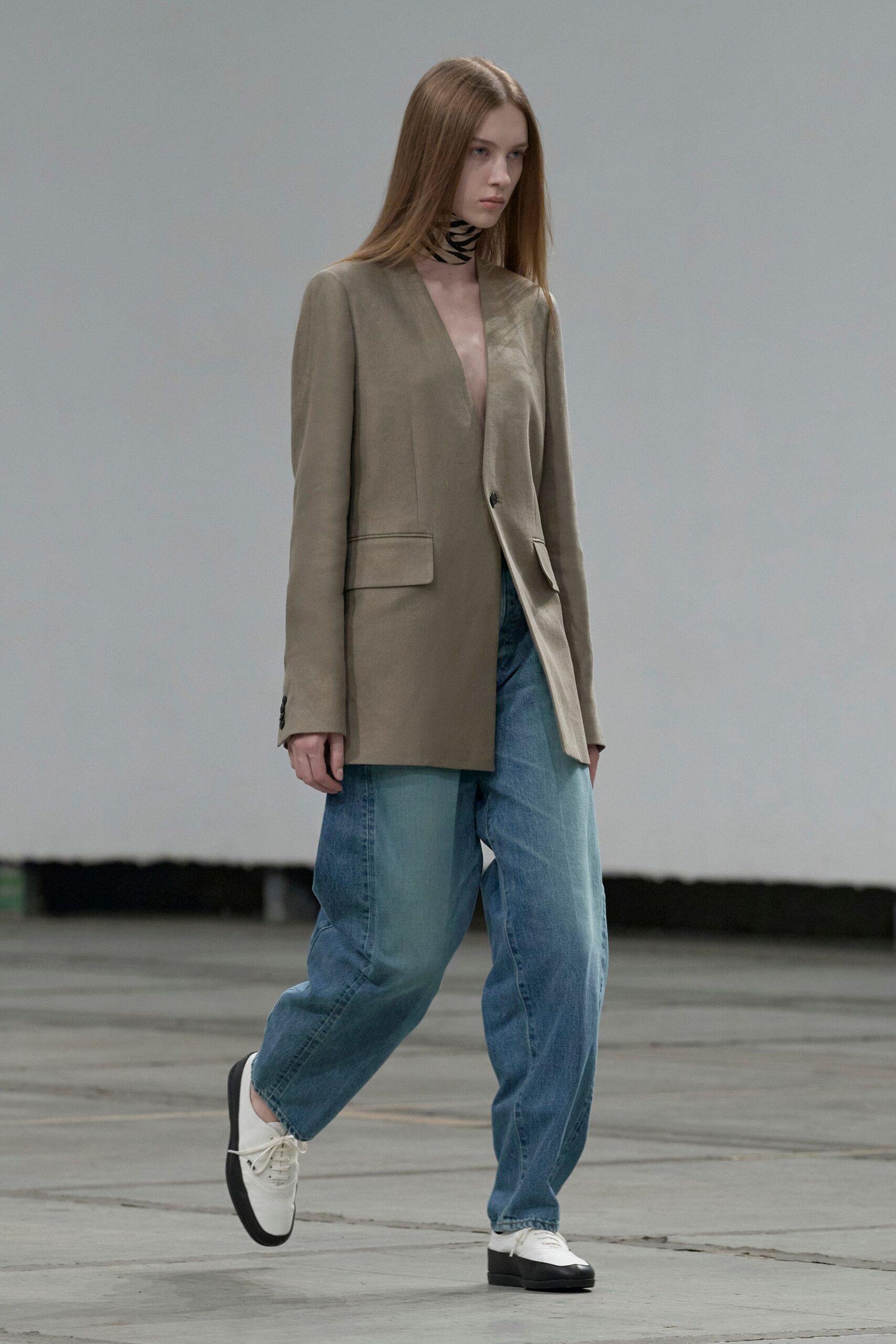Cream neck scarf ringed with uneven black stripes. Taupe collarless blazer in dry suiting, cut to a plunging single-button front. Pale blue jeans ballooning through the leg with a soft stack at the hem. White derbies with blunt black dipped soles. Crossing an empty convention hall before the booths are built.