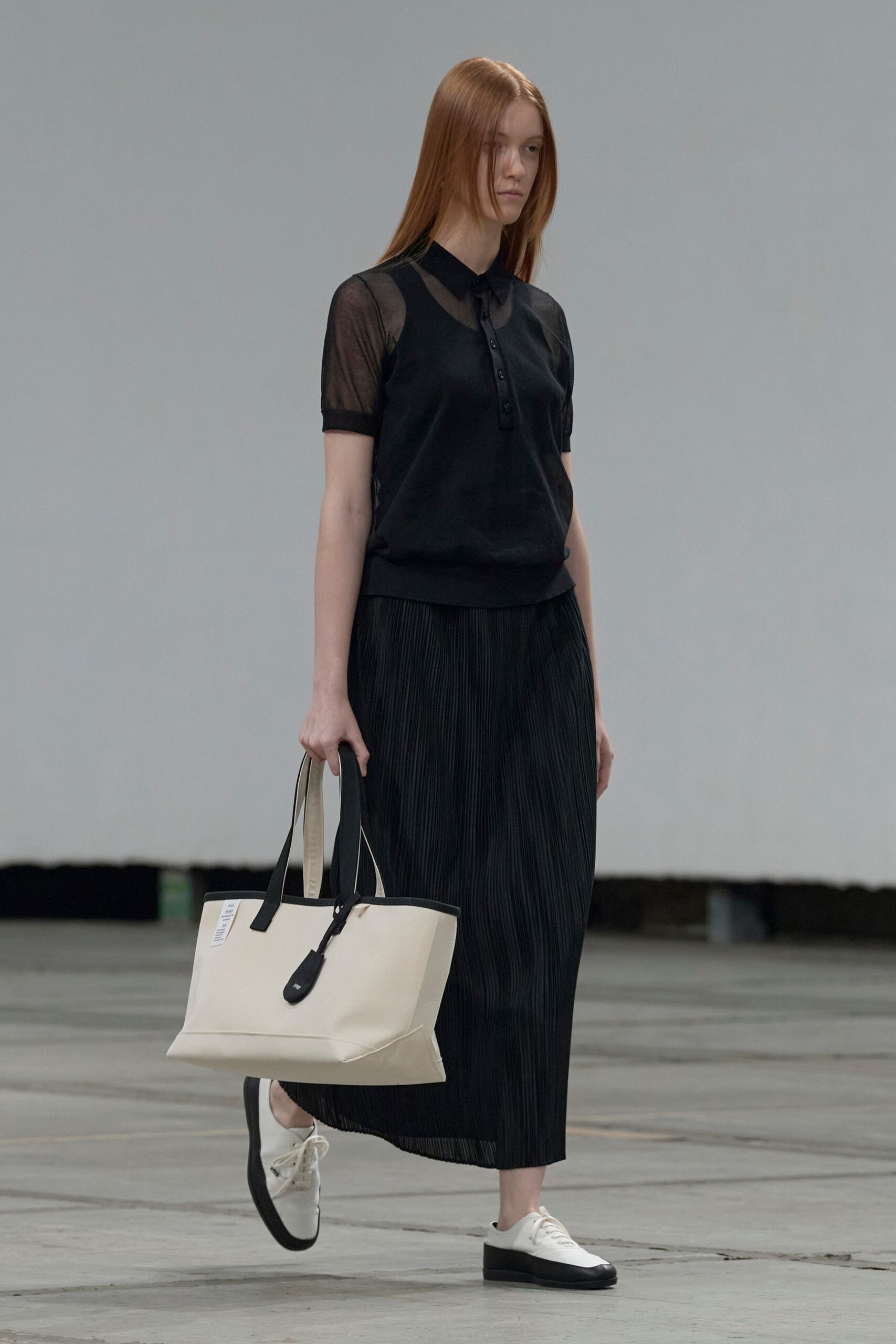 Black polo top with sheer mesh sleeves and a narrow button placket. Black midi skirt in fine vertical pleats, hem hanging slightly translucent. Cream canvas tote with black piping and a long dangling zip tab. White derbies on thick black rubber soles. Crossing an echoing concourse before the first commuter train fills it.