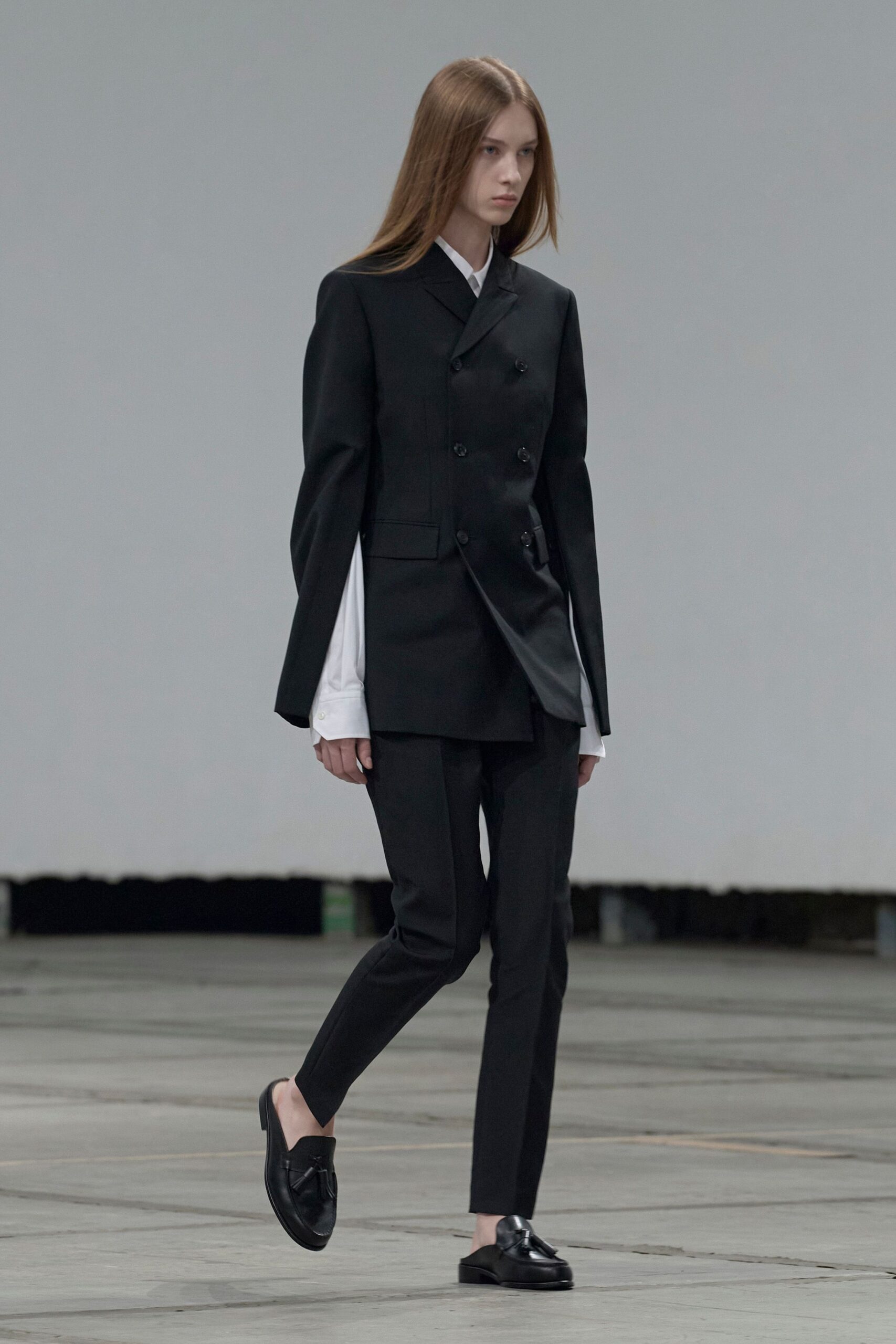 Black double-breasted blazer with slit sleeves and a skewed hem overlap. White poplin shirt, cuffs dropped long below the jacket sleeves. Slim black trousers with a sharp front crease. Black leather backless loafers with tassels at the vamp. Early train to a tribunal hearing, coffee cooling in your hand.