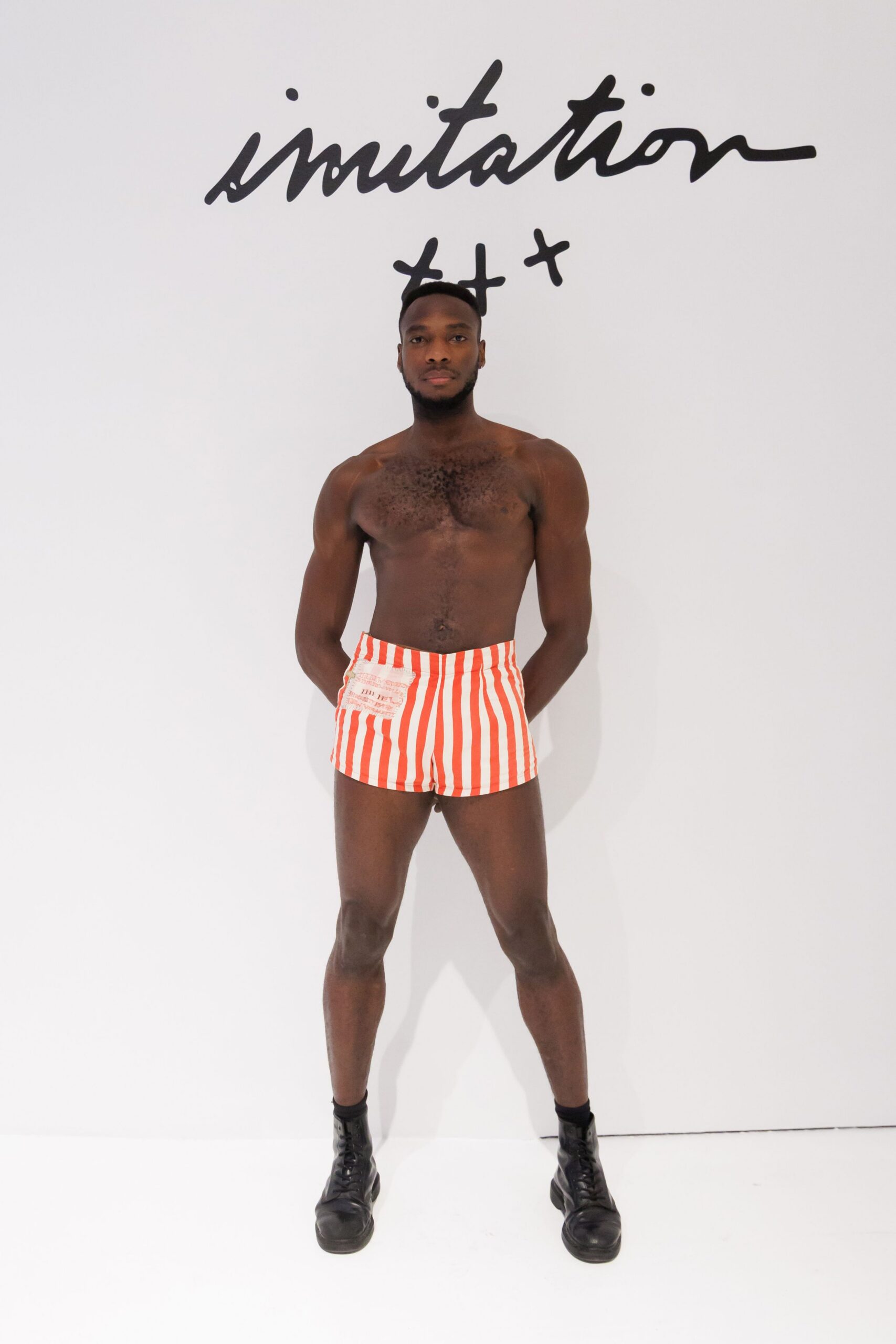 Red-and-white vertical striped shorts with a raw-edged patch pocket. Black leather combat boots with thick rounded toes. Boardwalk changing room, towel still damp and sand on the floor.