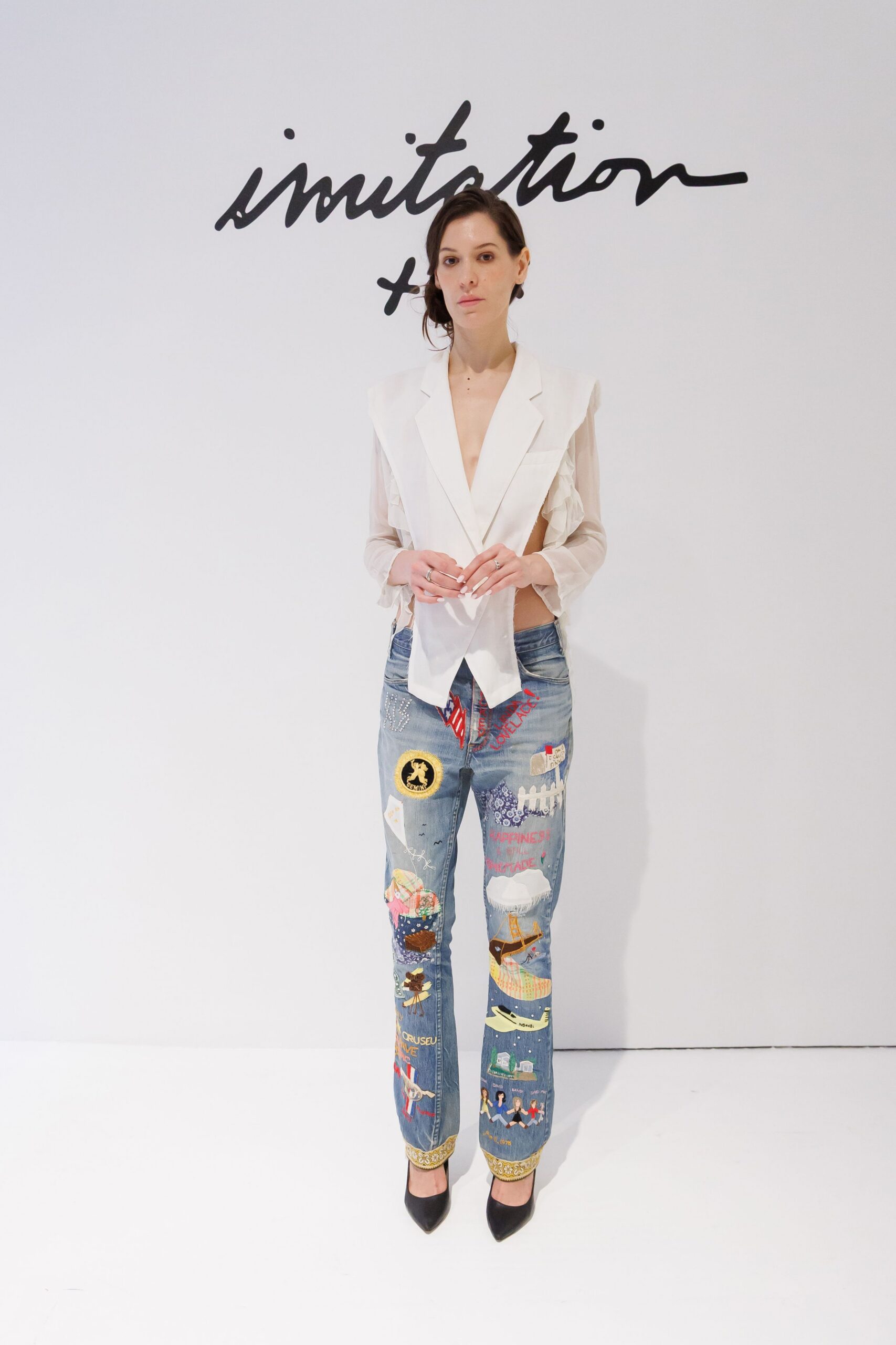 Ivory sheer blouse cut like a vest, plunging lapels framing bare sides. Light-wash jeans crowded with stitched storybook patches and a gilt trouser hem. Black pointed pumps. For reading old postcards in a seaside flat after the rain stops.