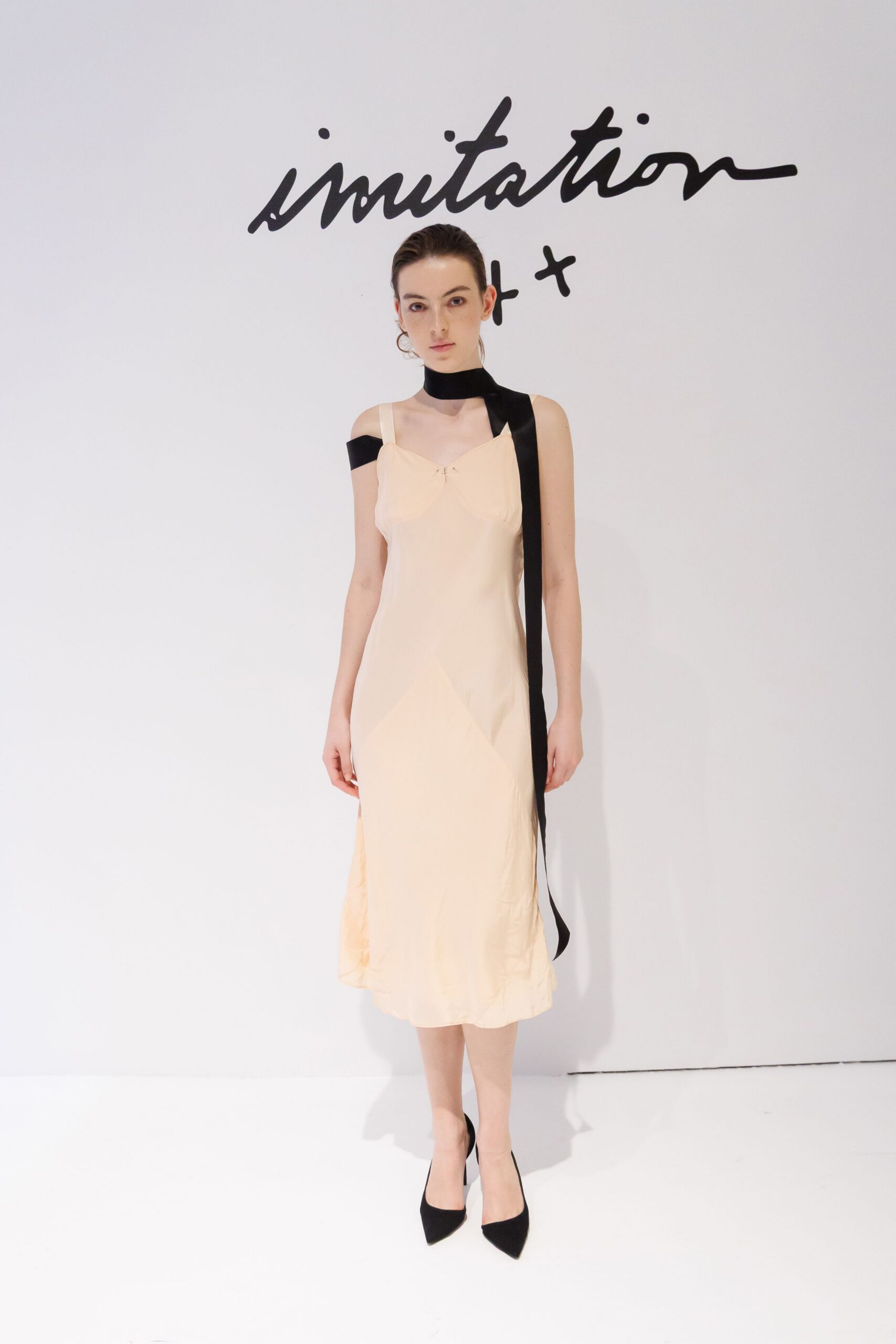 Black satin scarf looped at the neck, one end falling past the knee. Peach bias-cut slip dress with tiny tack stitches at the bust. Black pointed pumps in matte suede. For standing in a hotel corridor after midnight, keycard gone missing.