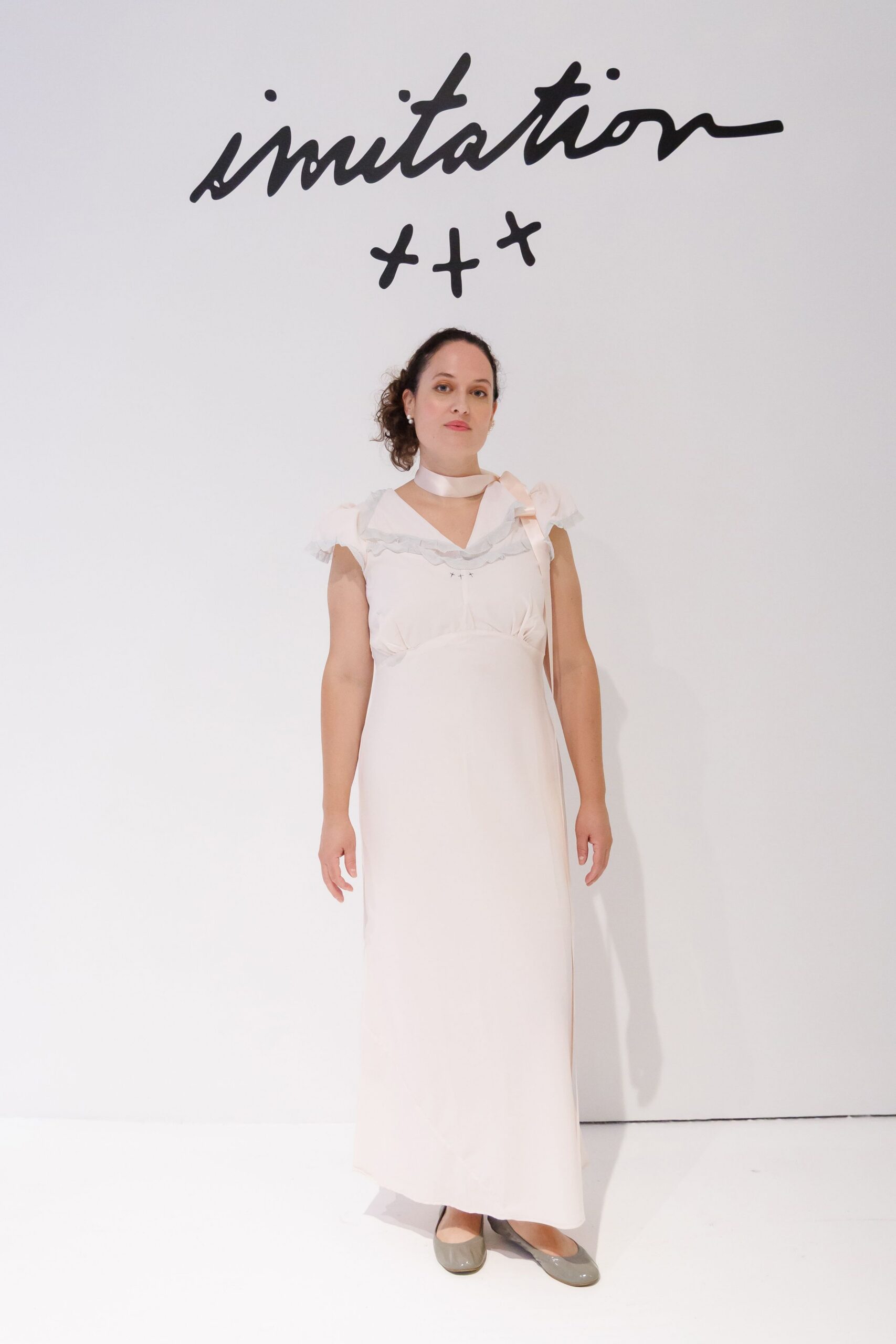Pale satin neck scarf tied in a loose side bow. Blush floor-length dress with flutter sleeves and a rippled ruffle neckline. Grey patent square-toe flats. Registry office lobby, flowers sweating in brown paper by your feet.