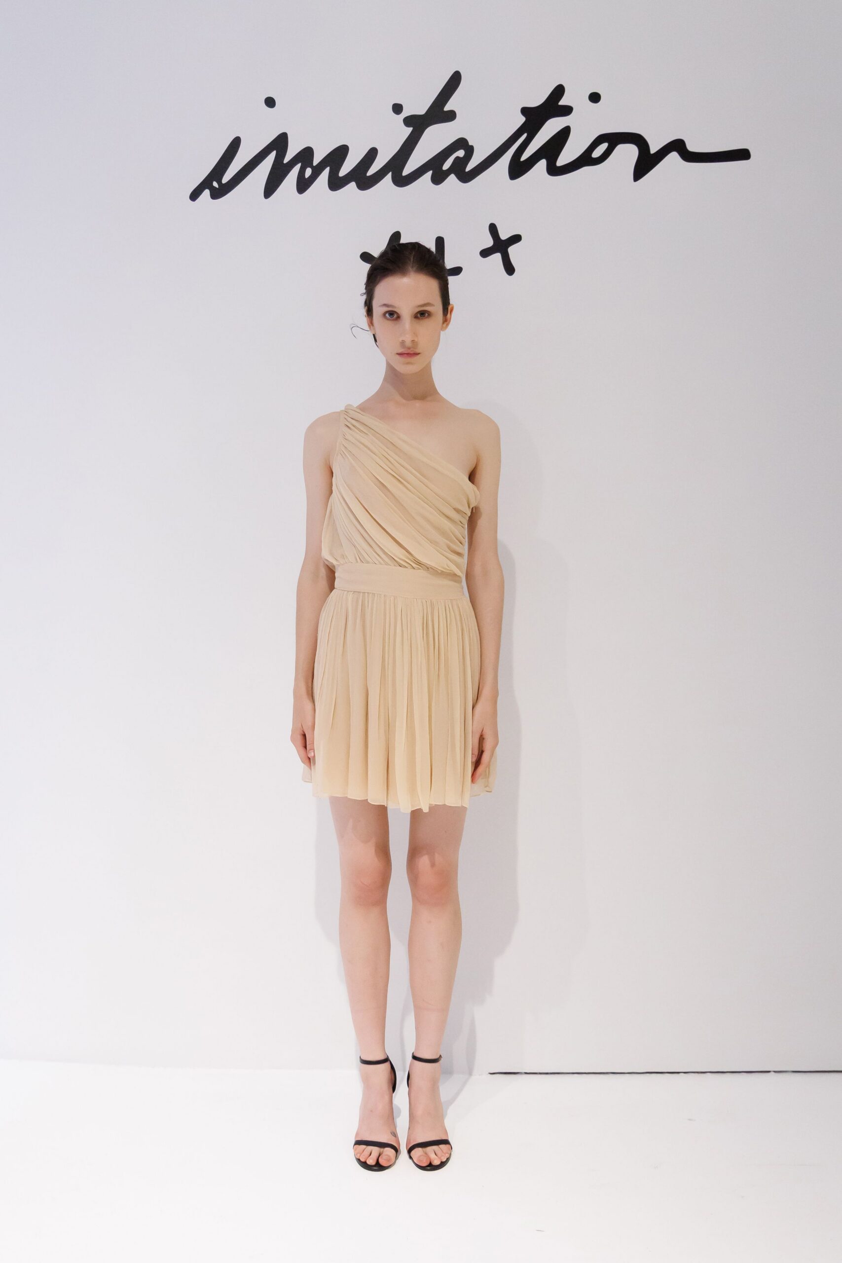 Pale beige chiffon mini dress, one-shoulder bodice densely ruched on the diagonal. Black ankle-strap sandals with bare, fine straps. For standing in a quiet hotel lobby before someone else's wedding.