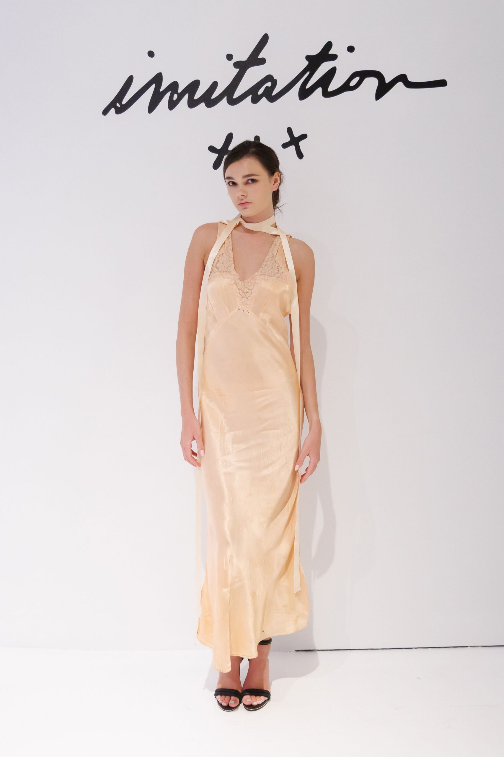 Peach satin slip dress with lace inset at the deep V. Long ivory ribbons looped at the throat, ends hanging past the hips. Black slide sandals with a squared, barely there strap. Morning after a hotel wedding, still wearing the room slippers downstairs.