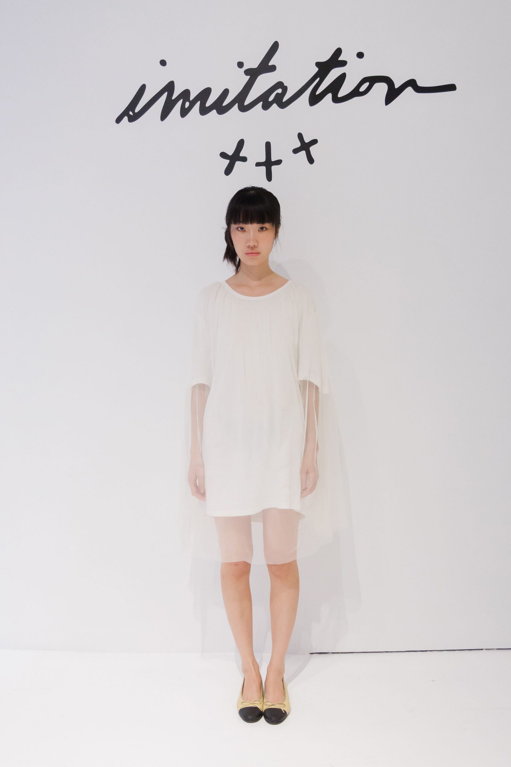 Ivory shift dress with a sheer tulle overskirt falling past the knees. Cream cap-toe pumps, tiny bow pinched at each vamp. Standing in a rental gallery before the folding chairs are set out.