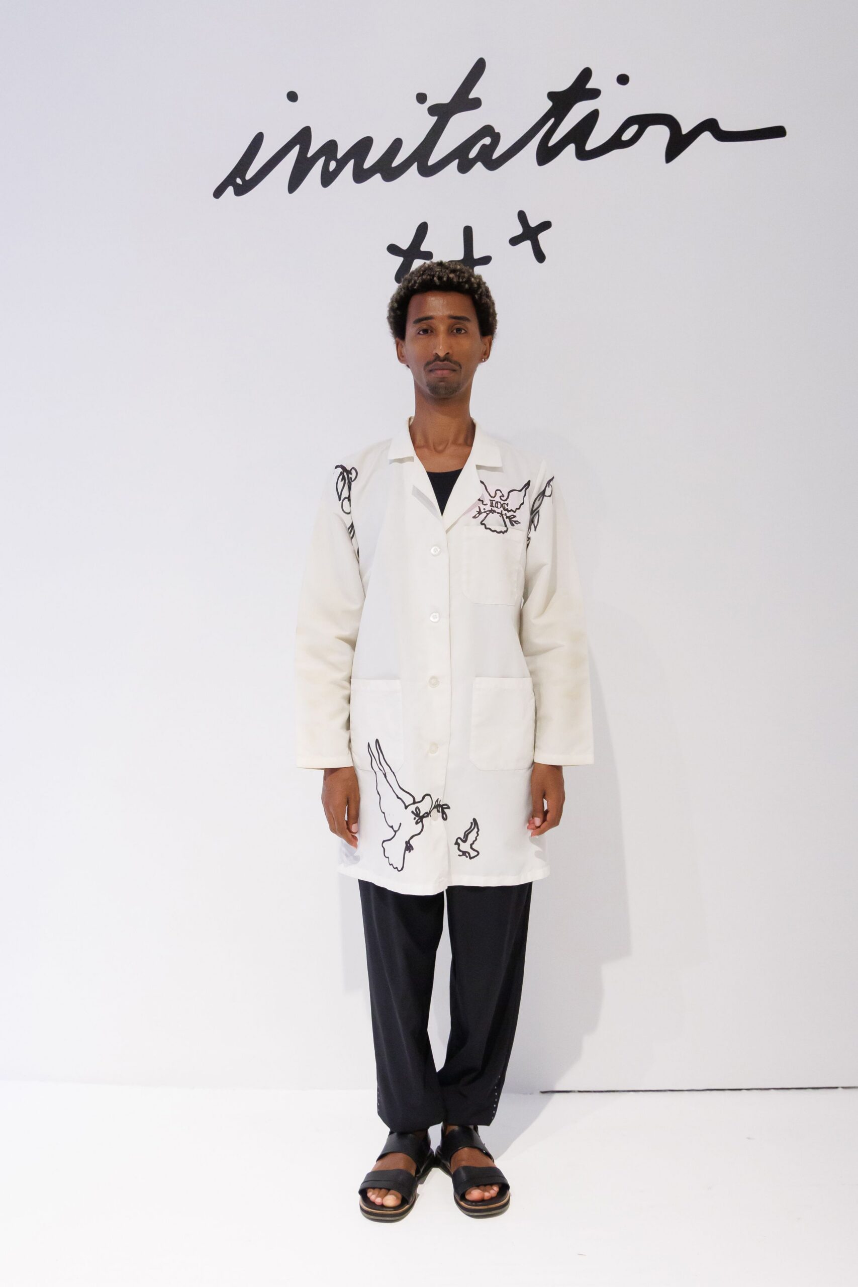 White workwear coat with black line-drawn birds skimming the hem. Black scoop-neck layer peeking through the open camp collar. Loose black trousers with a straight fall over the instep. Black slide sandals on a thick striped sole. For unlocking the gallery early, mop bucket still drying by the door.