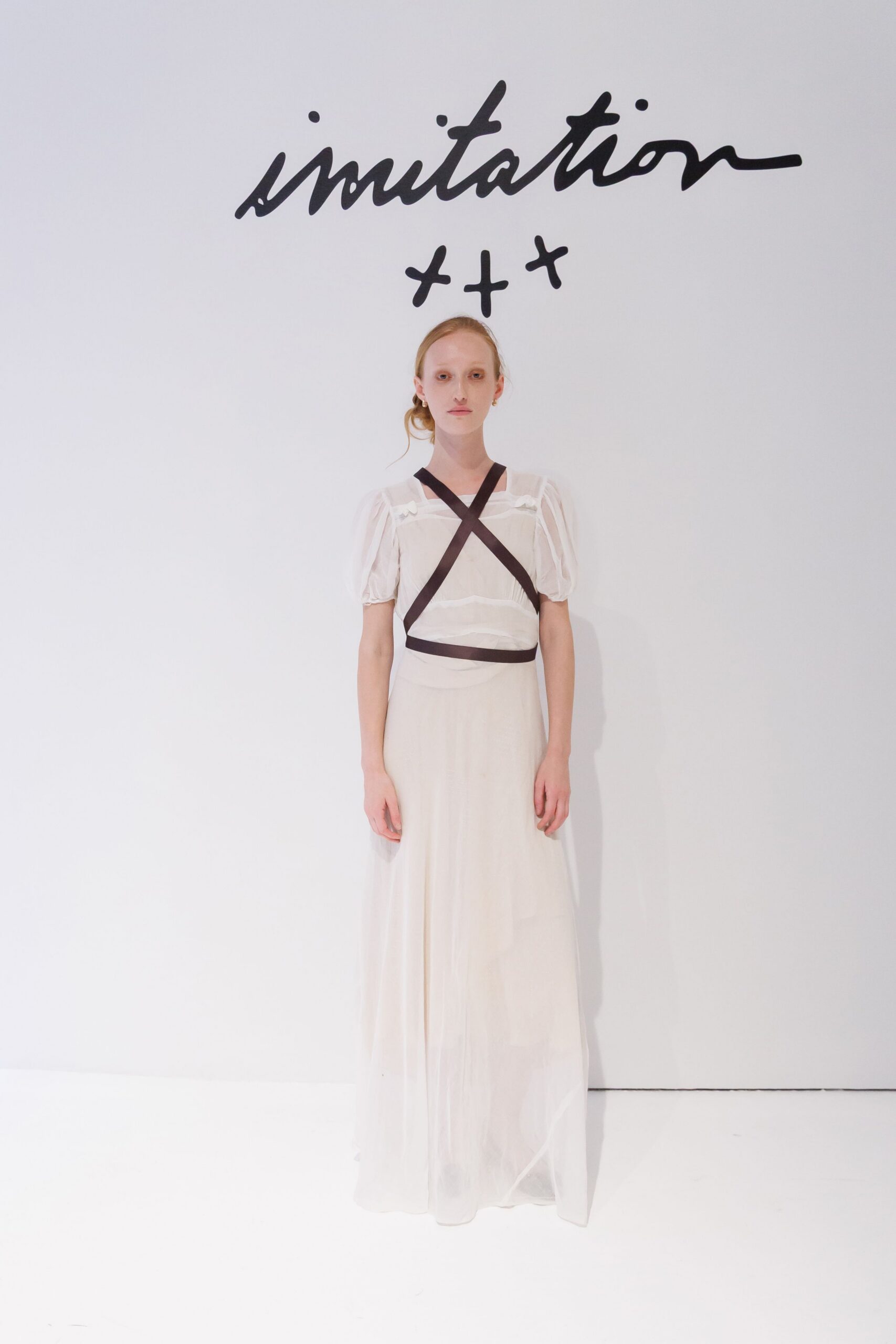 Ivory gauze dress with puff sleeves and a floor-drifting sheer skirt. Black grosgrain straps crossing the chest, cinched in a narrow waist band. Tiny gold hoops. Church hall quiet after the folding chairs have been stacked.