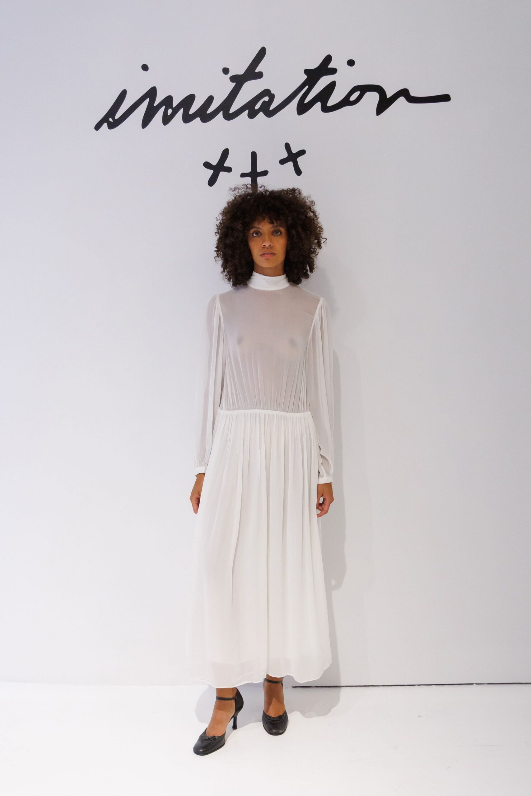 Sheer white georgette dress with a high neck and fine vertical pleats. Black Mary Jane heels with slim straps across the instep. Morning civil ceremony, paper forms waiting on a laminate table.