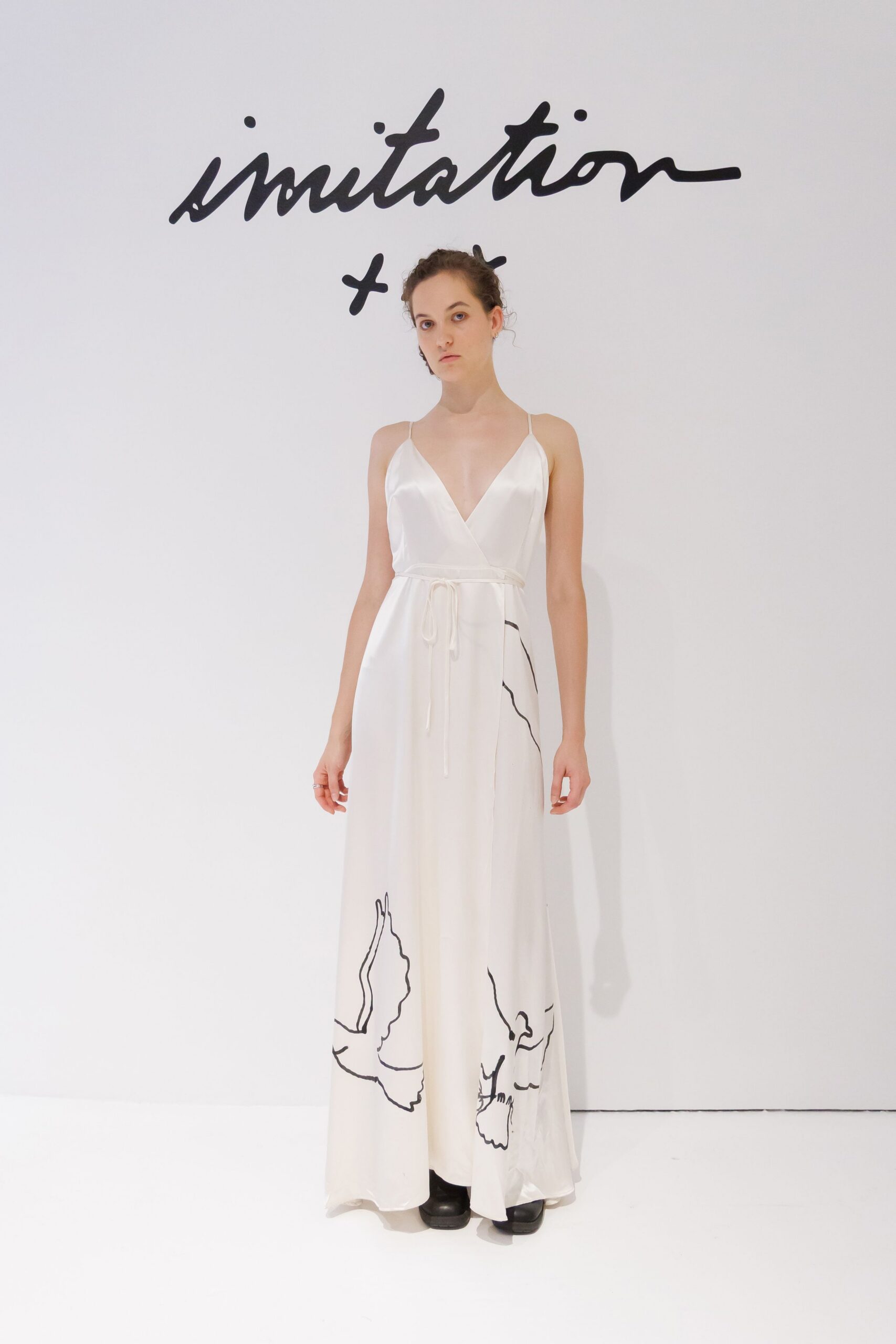 Ivory satin slip dress with a deep wrap V and cord-tied waist. Black platform shoes peeking from the pooling hem. Tiny dark drop earrings. Morning of the civil ceremony, flowers still wrapped in paper.