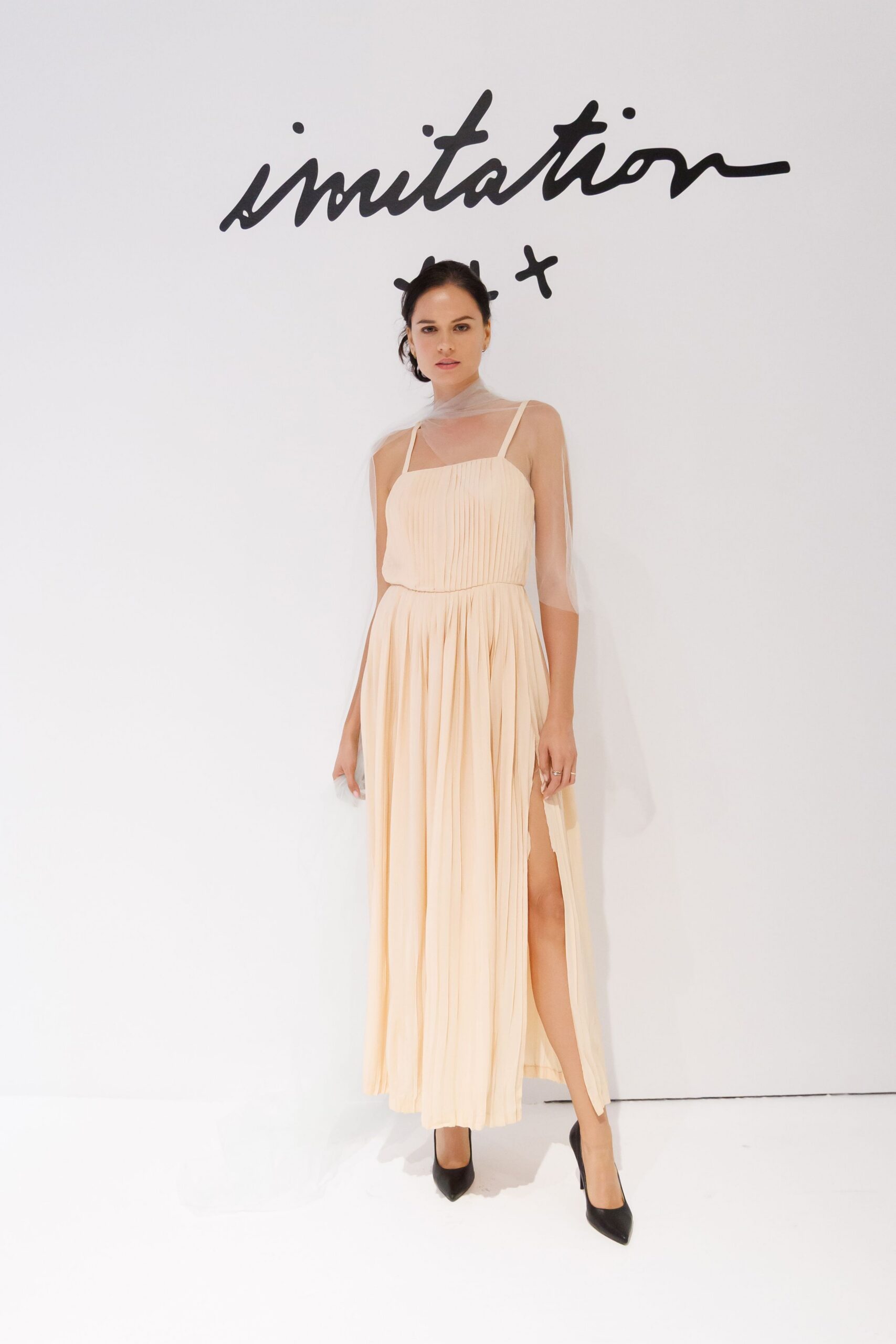 Sheer tulle scarf looped once at the throat, tails falling past the elbows. Blush chiffon slip dress with knife-pleated bodice, thigh slit breaking the skirt. Black pointed pumps. For the courthouse wedding where someone brought supermarket flowers.