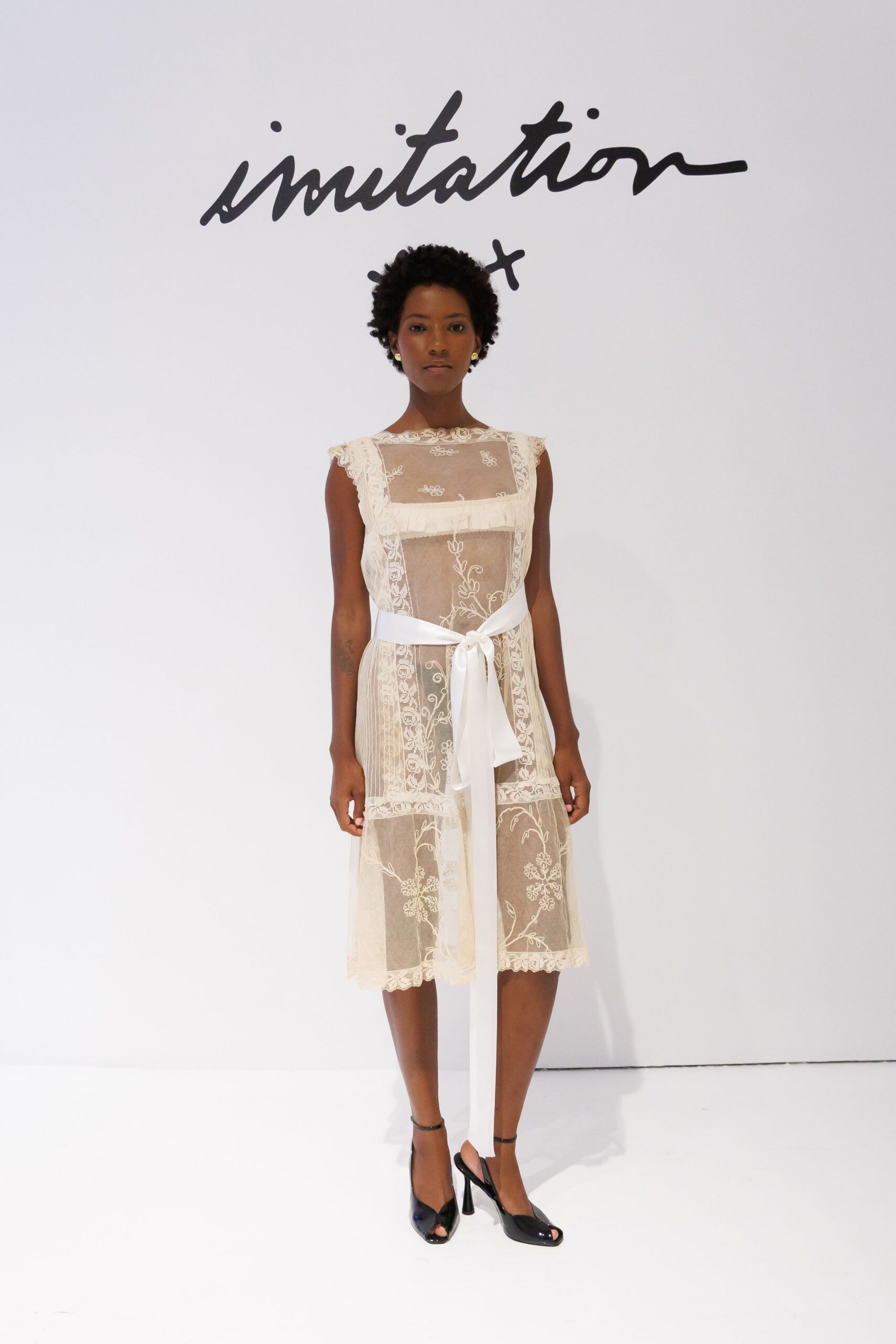 Sheer ivory lace dress with floral embroidery and a square illusion yoke. Wide satin sash tied low, one tail dropping past the hem. Black patent slingback heels with peep toes. Registry office stairs, bouquet paper already soft from your hands.