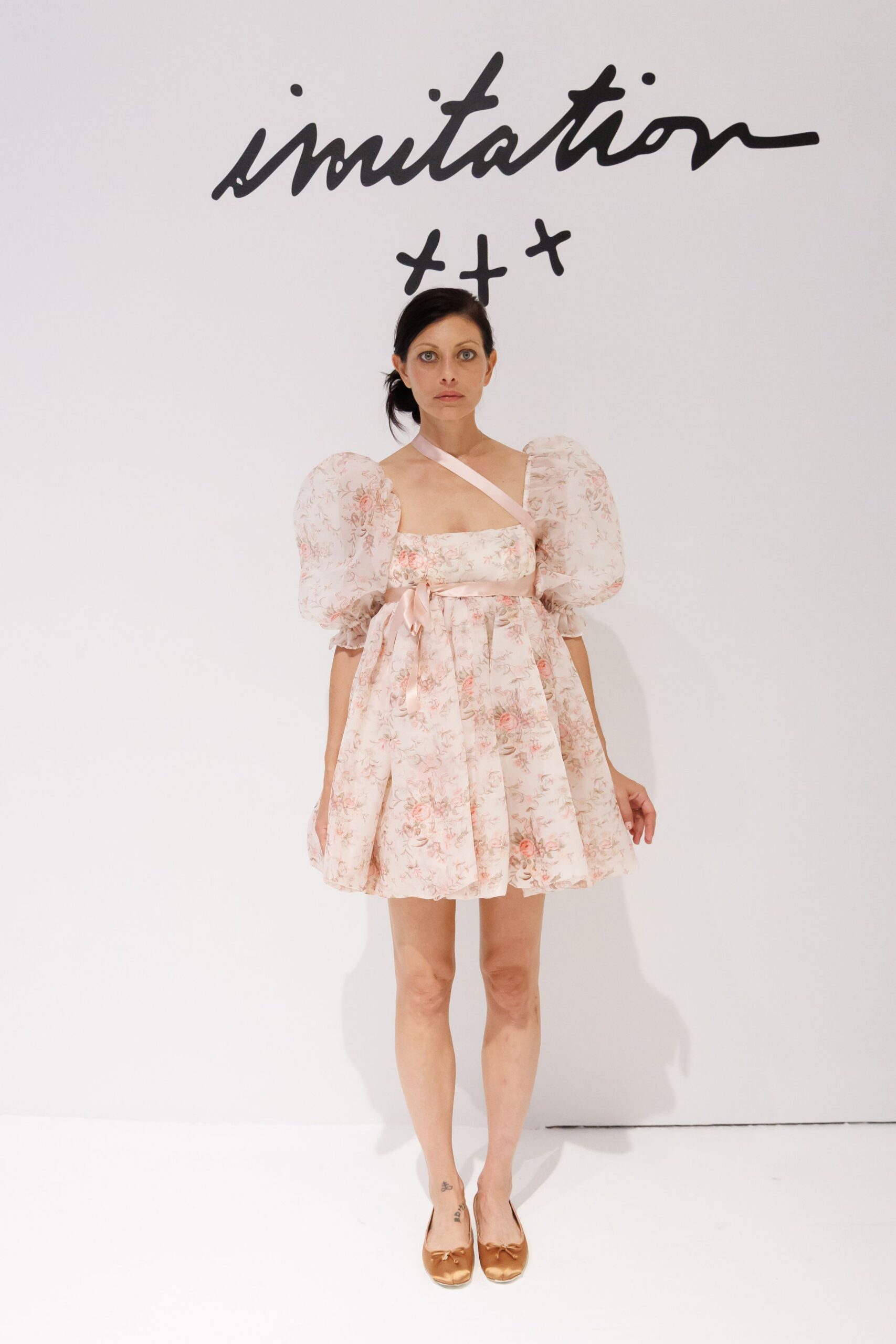 Blush floral mini dress with puffed organza sleeves and a satin shoulder strap. Bronze satin ballet flats with tiny bow knots at the square toe. Garden wedding reception, cake plates stacked beside the melted candles.