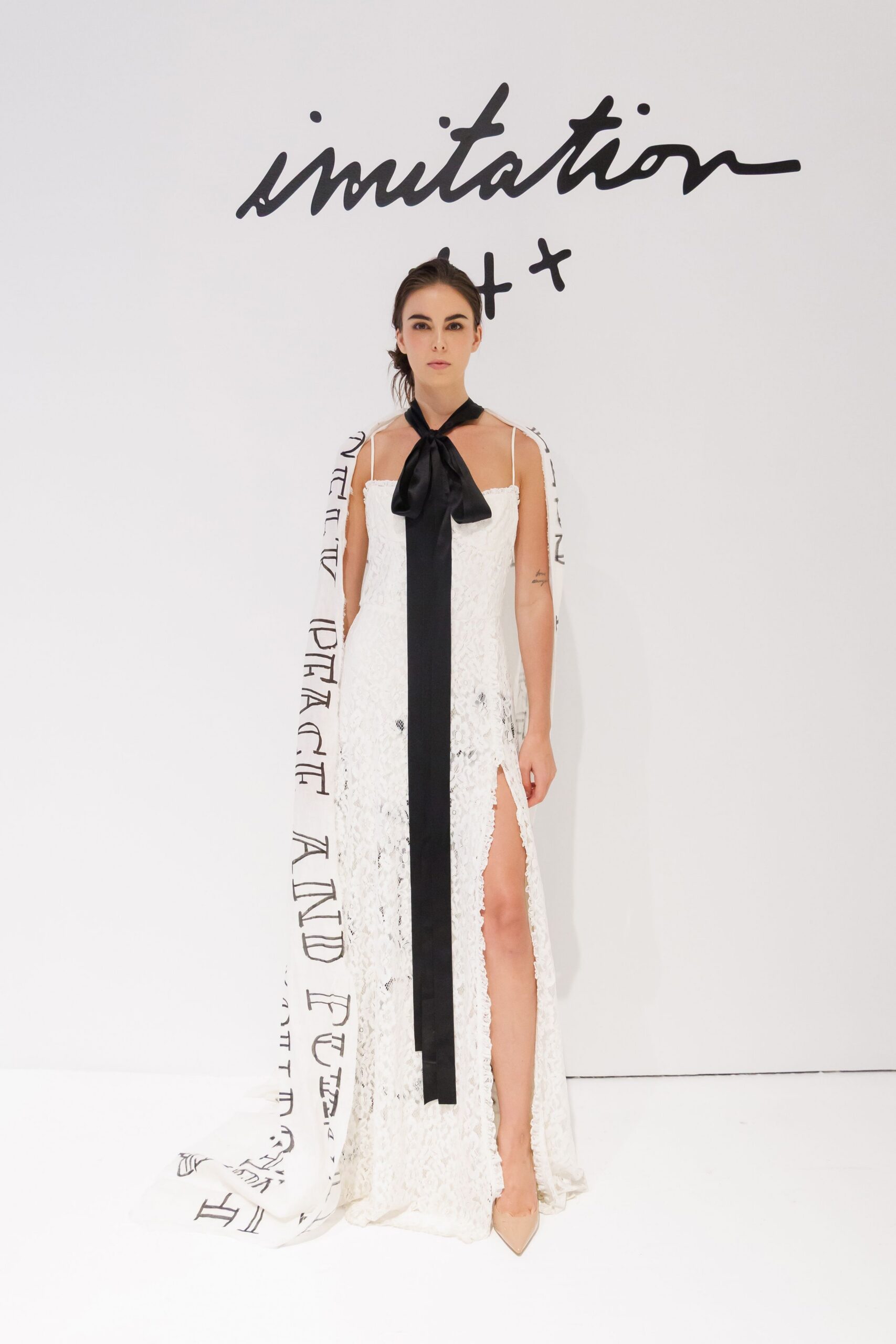 Black satin neck scarf tied in a broad drooping bow. White lace column gown with thin straps and a high thigh slit. Long white train printed with black lettering, pooling off to one side. Nude pointed pumps. For reading vows in a municipal hall with the florist stuck in traffic.