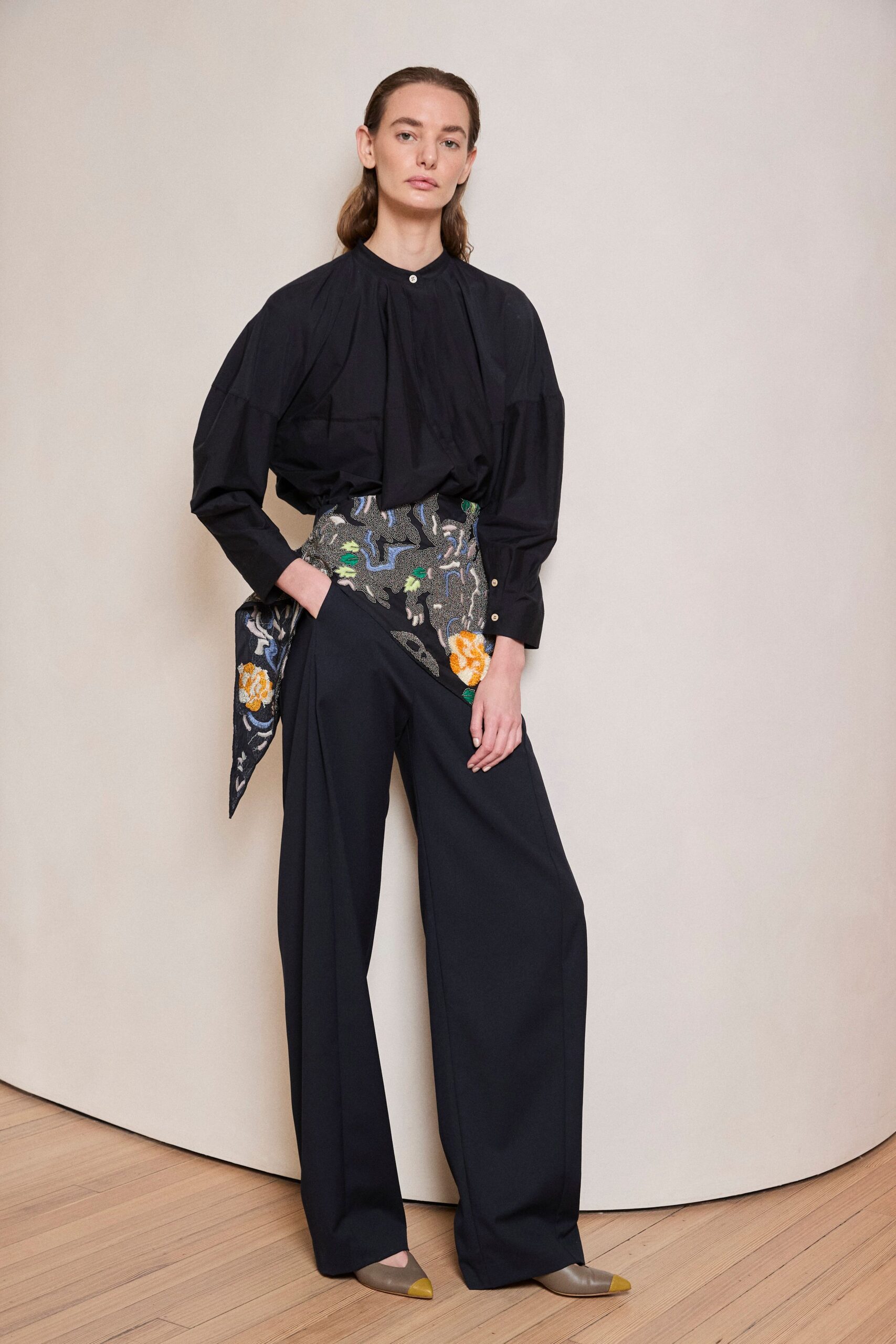 Black cotton blouse with a stand collar and gathered front fall. High-waisted black wide-legs cut with a sweeping embroidered wrap panel. Matching tapestry scarf hanging from the hip, one point sharpened. Taupe leather flats with mustard cap toes. For opening a door onto a quiet apartment dinner above the river.