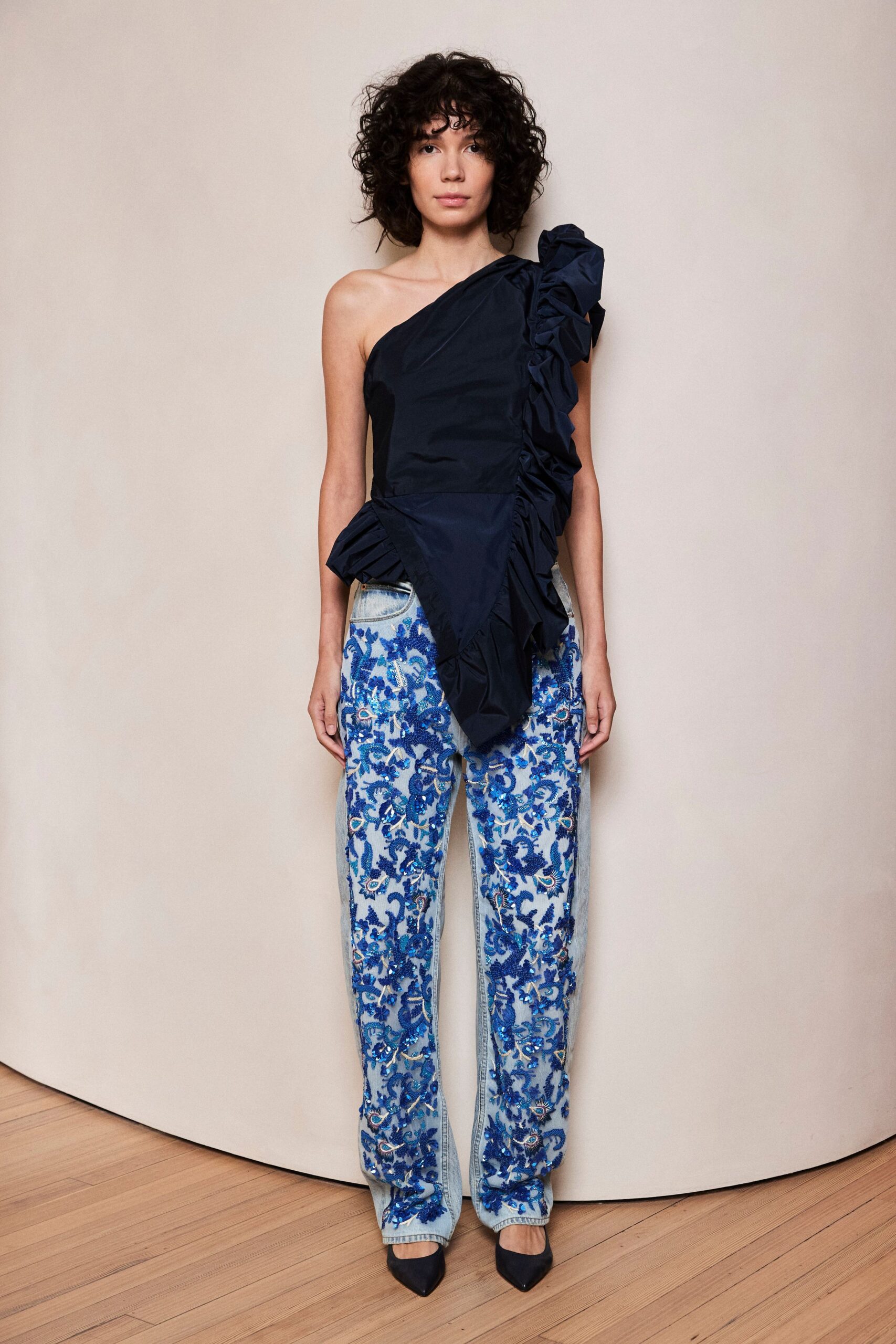 Midnight taffeta one-shoulder top with a densely ruched cascading ruffle. Pale denim jeans overlaid in cobalt beaded vine embroidery, pockets left bare. Dark satin pointed pumps. For arriving at a garden wedding after the grass has gone damp.
