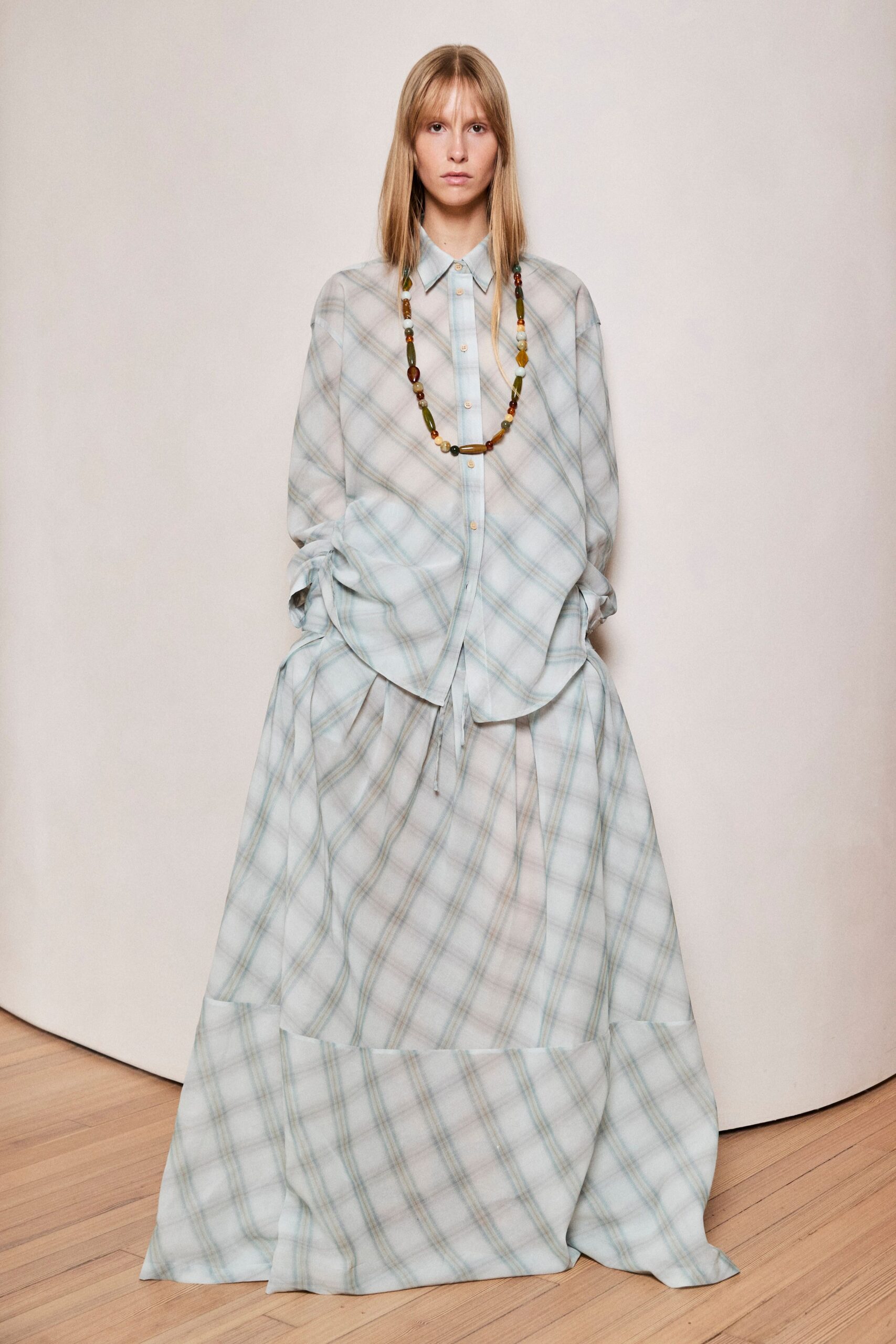 Long strand of mottled resin beads in olive, amber and milk. Powder-blue plaid shirt in gauzy cloth, hem curling into soft points. Matching full skirt with a drawstring waist and deep folded hem. Sunday house tour, socks sliding on waxed floorboards.