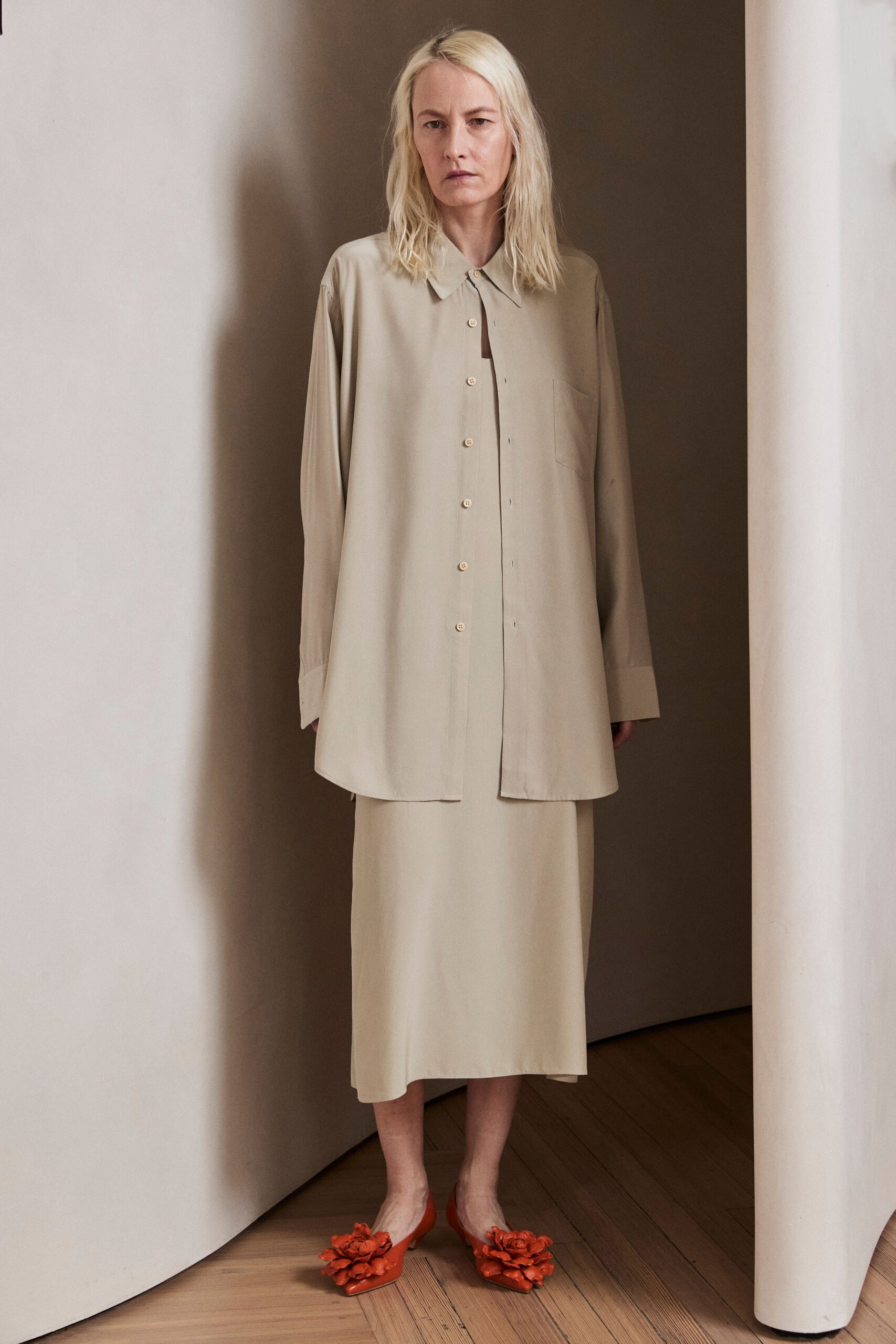 Long bone shirt in fluid matte cloth, one button left undone. Cream column skirt falling straight from under the shirttail. Burnt orange pointed mules topped with oversized leather flowers. For crossing a silent hotel corridor before anyone else is awake.