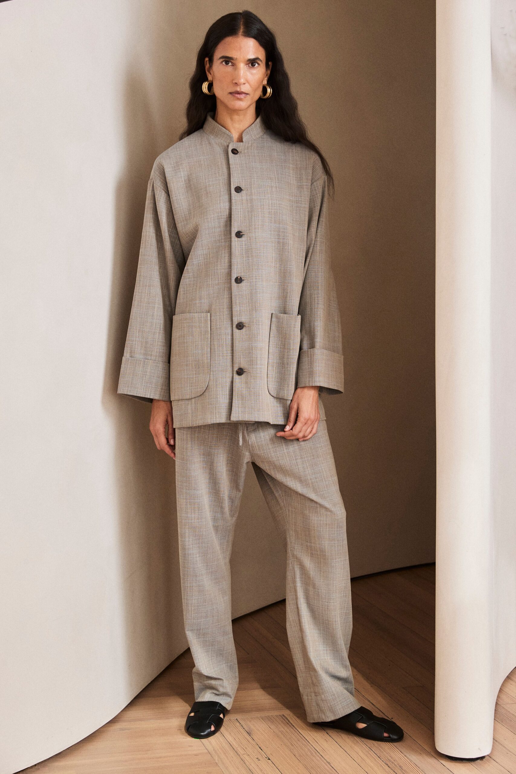 Thick gold hoops hugging the lobe. Greige slub-weave collarless jacket, dark horn buttons and rounded patch pockets. Matching straight trousers with a white drawcord peeking at the waist. Black cutout leather slides. For the first morning in a rental house where nobody has unpacked yet.