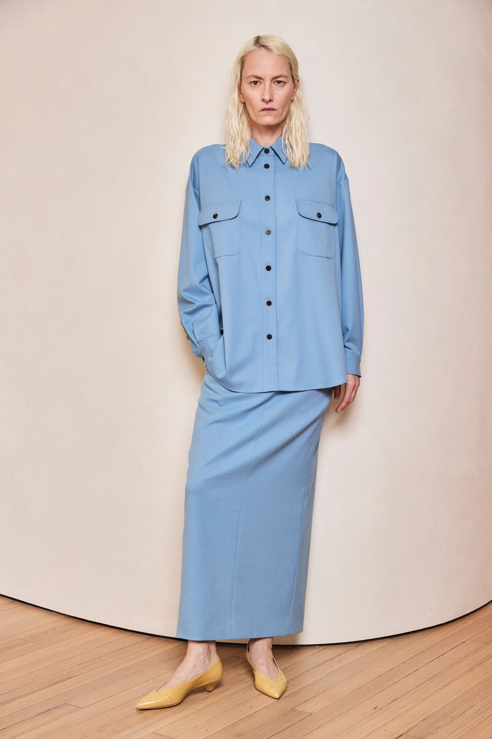 Powder-blue fluid shirt with oversized flap pockets and black buttons. Matching column skirt, center seam running straight to the shin. Butter-yellow pumps in glossy patent leather. Early meeting in a glass office before the heat has lifted.