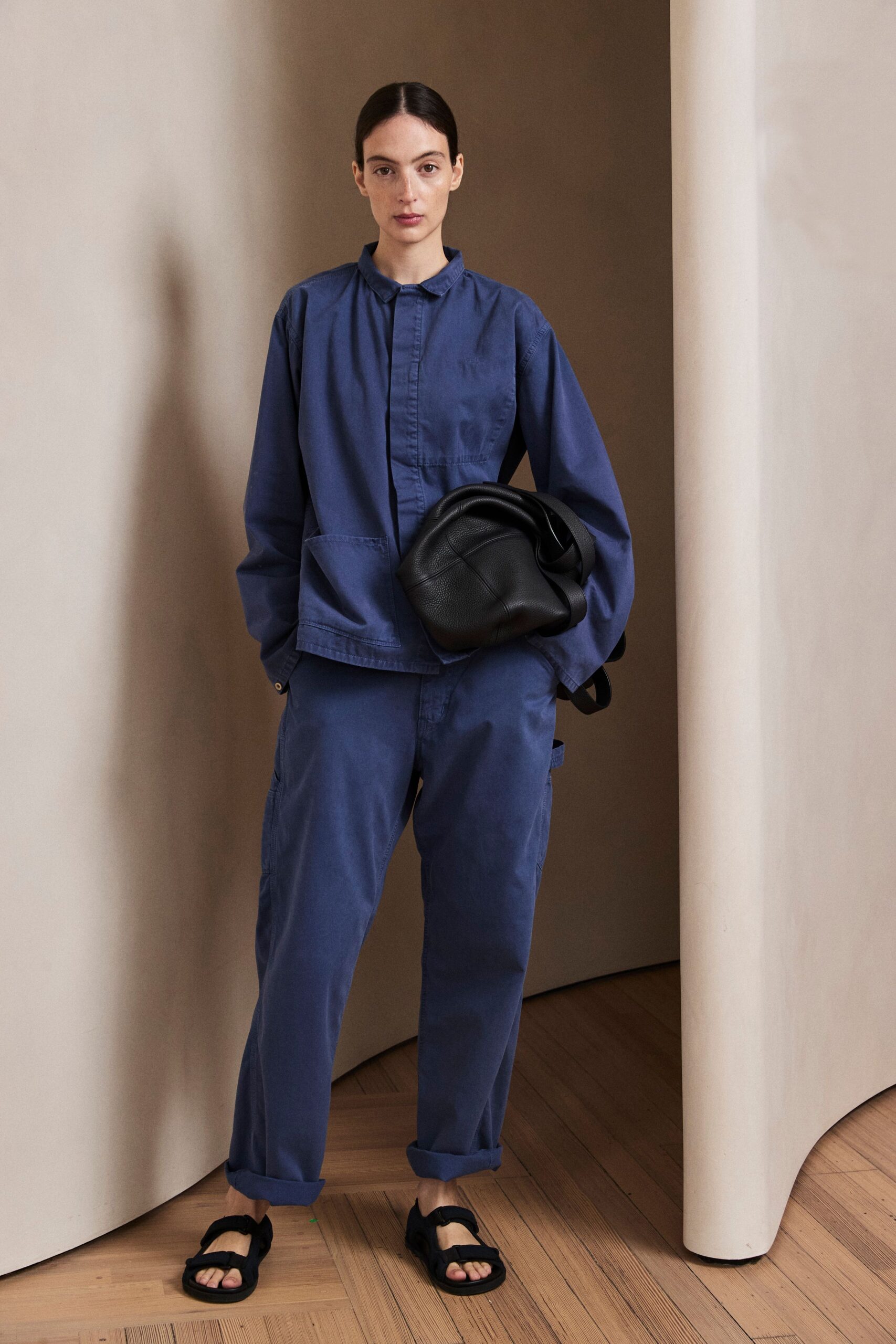 Washed indigo cotton shirt jacket with concealed placket and skewed patch pockets. Matching indigo twill cargos, loose through the leg with turned cuffs. Slouchy black pebbled leather bag, folded into a soft crescent. Black sandals with thick velcro straps. For slipping out to buy bread before the street fully wakes.