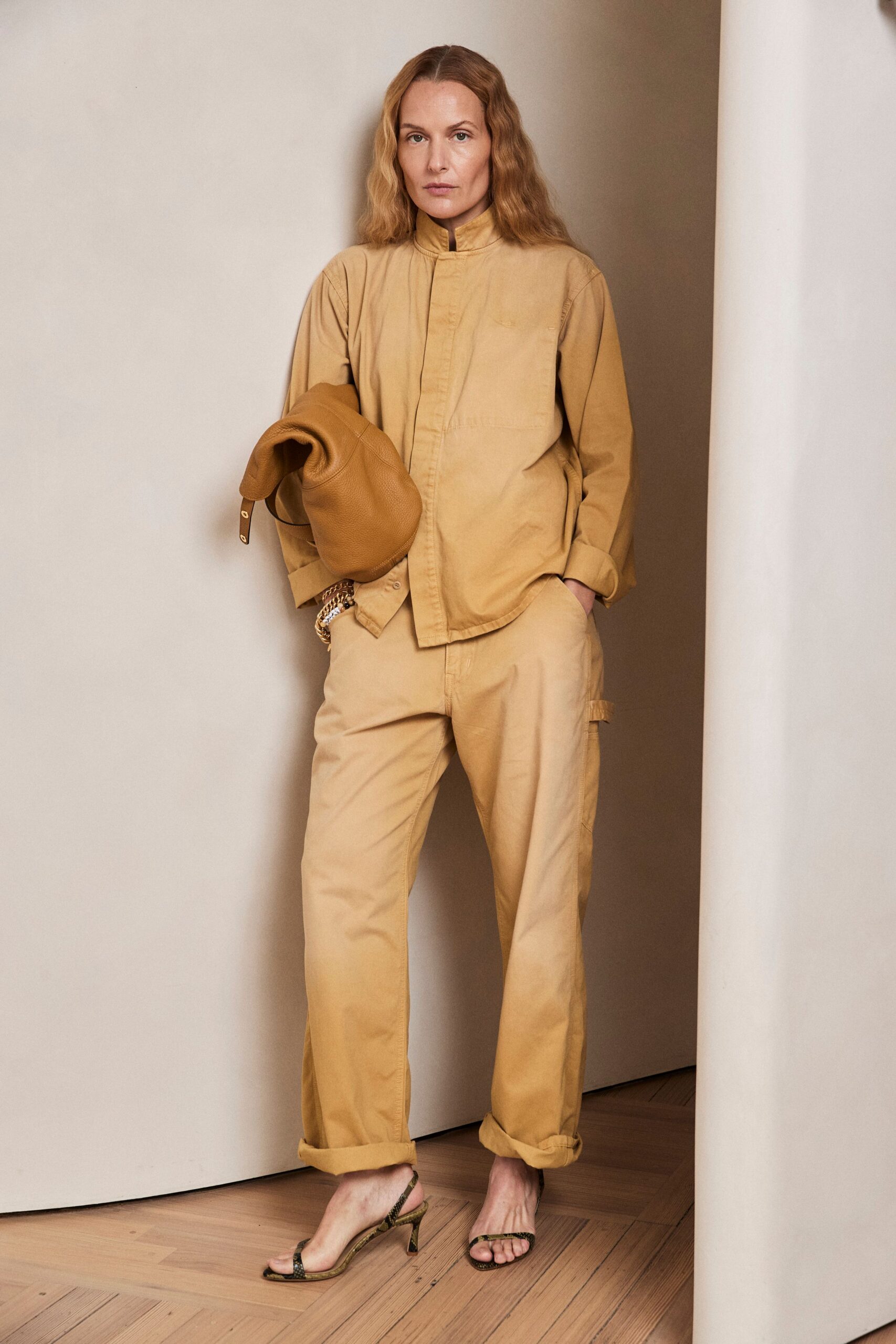 Faded ochre cotton shirt with a stand collar and hidden placket. Matching straight-leg cotton cargos, cuffs rolled thick above the ankle. Slouchy pebbled leather clutch tucked under one arm. Python slingback sandals on a narrow kitten heel. For checking into a desert hotel before the room is ready.