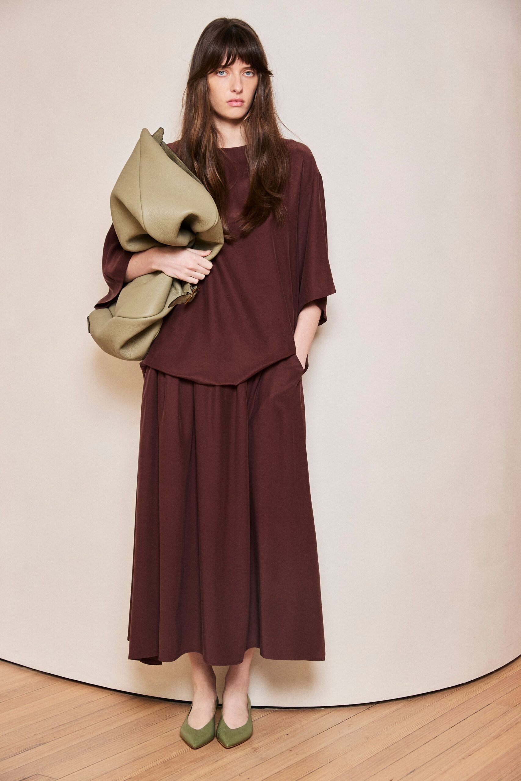 Burgundy crepe top with dropped shoulders and a softly skewed hem. Matching wide culottes, deep front pleats opening from the waistband. Oversized sage pebbled-leather clutch, folded into a slumped twist. Pointed sage flats in smooth leather. For picking up groceries after the florist wrapped the stems in brown paper.