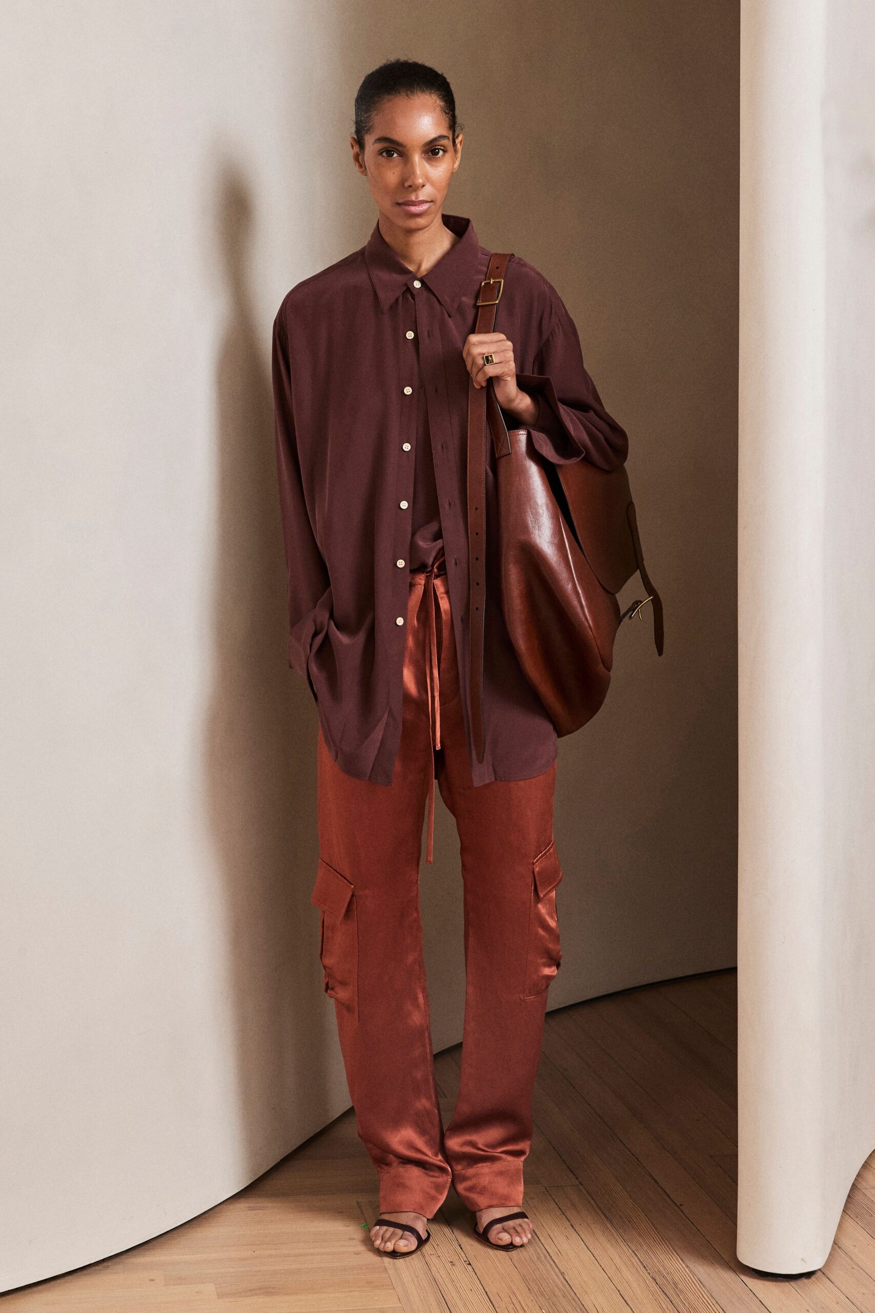 Oversized aubergine shirt in fluid matte crepe, ivory buttons marching off-center. Burnt copper satin cargo pants with flap pockets and a dangling drawcord. Cognac leather shoulder bag, slouching under a wide buckled strap. Dark brown thong sandals. For crossing a quiet hotel lobby before the conference coffee is set out.