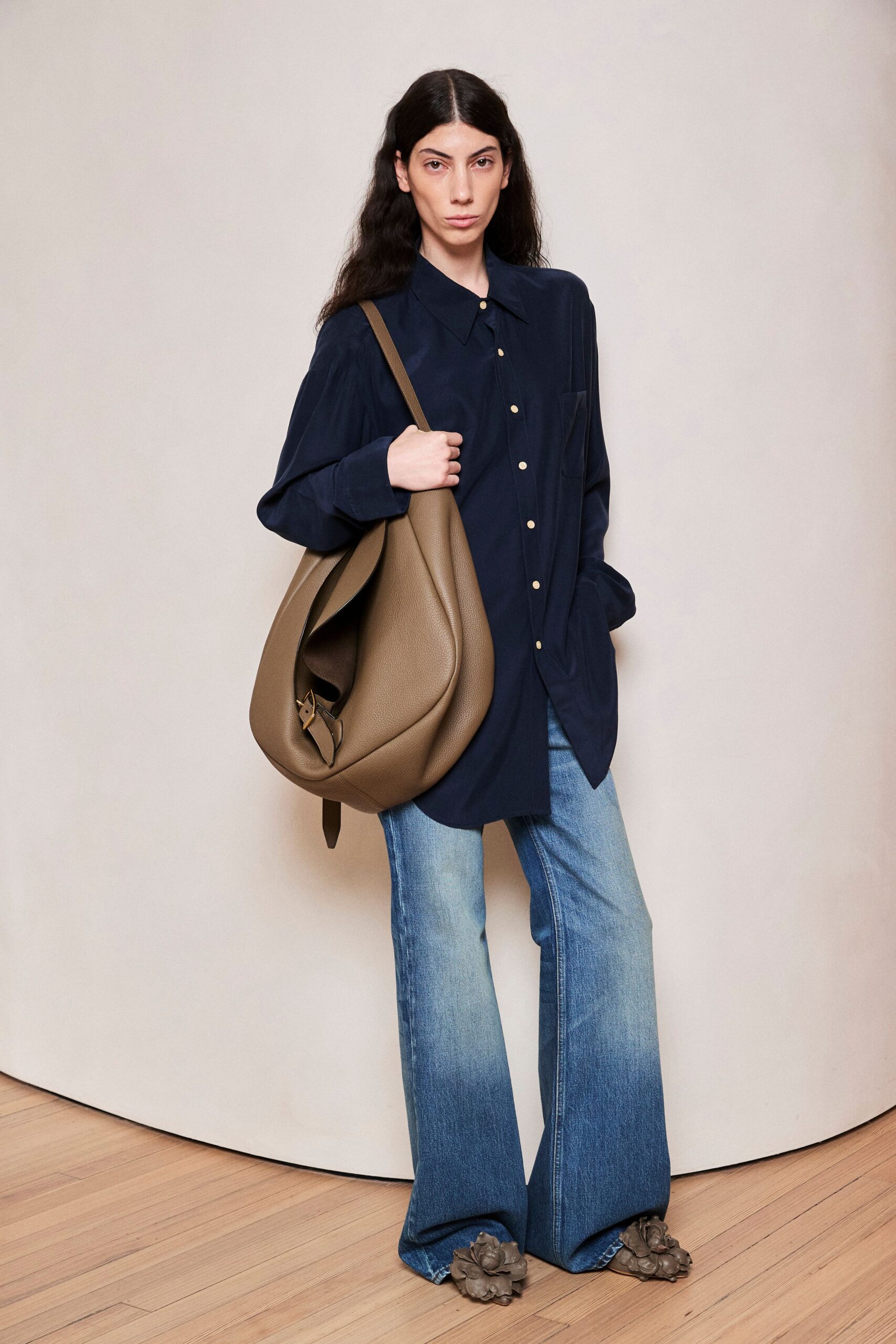 Taupe pebbled leather hobo slouched deep, strap buckle dangling low. Ink navy shirt hanging long, pale buttons marching off-center. Light-wash flared jeans, whiskered through the thigh and pooling. Leather shoes buried under oversized sculpted flower covers. For the grocery run that turns into an afternoon downtown.