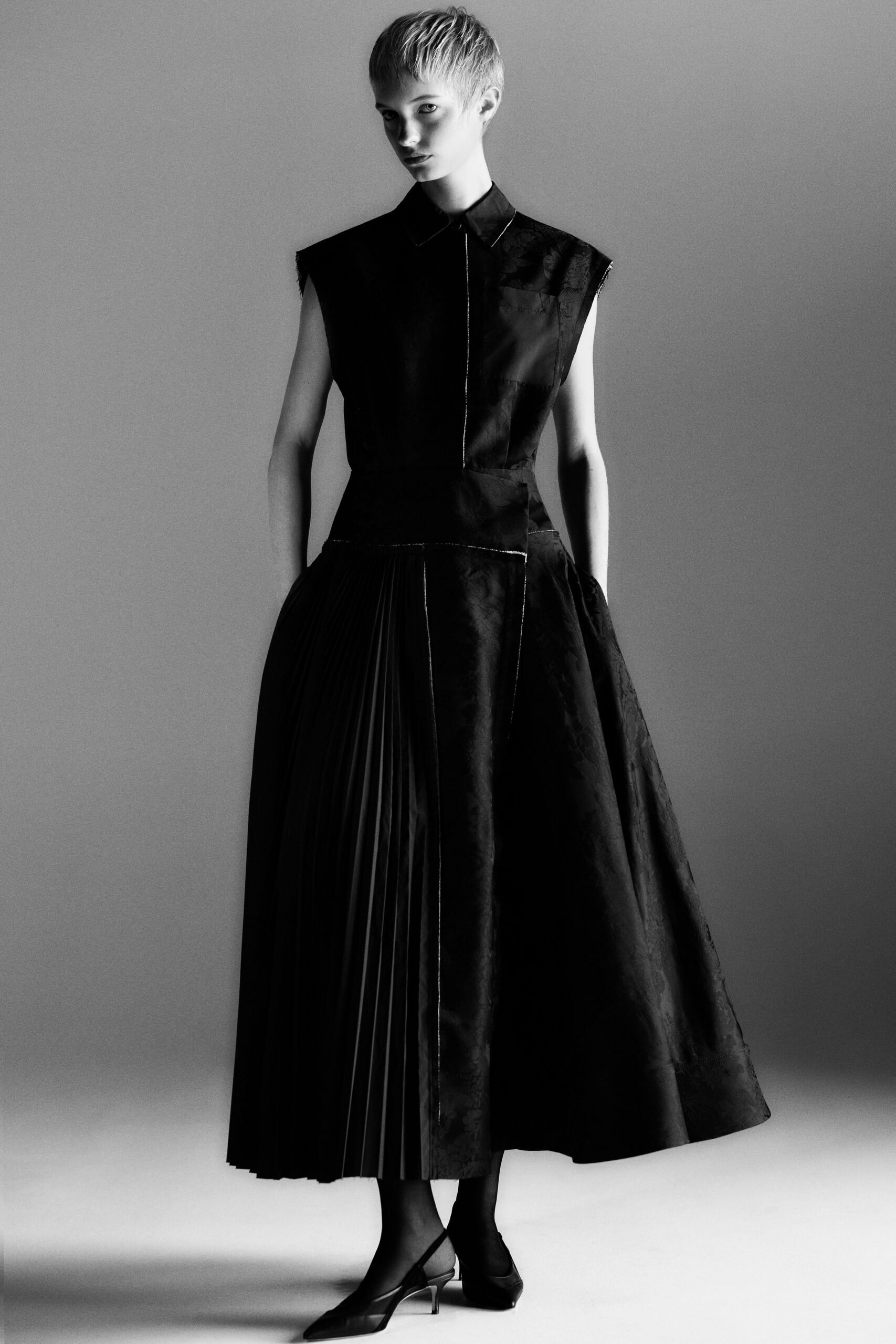 Black collared dress in mismatched matte and floral jacquard panels, one side finely pleated. Pointed mesh slingbacks with narrow kitten heels. Sheer black hosiery. Museum atrium at closing, footsteps carrying after the guards lock one wing.