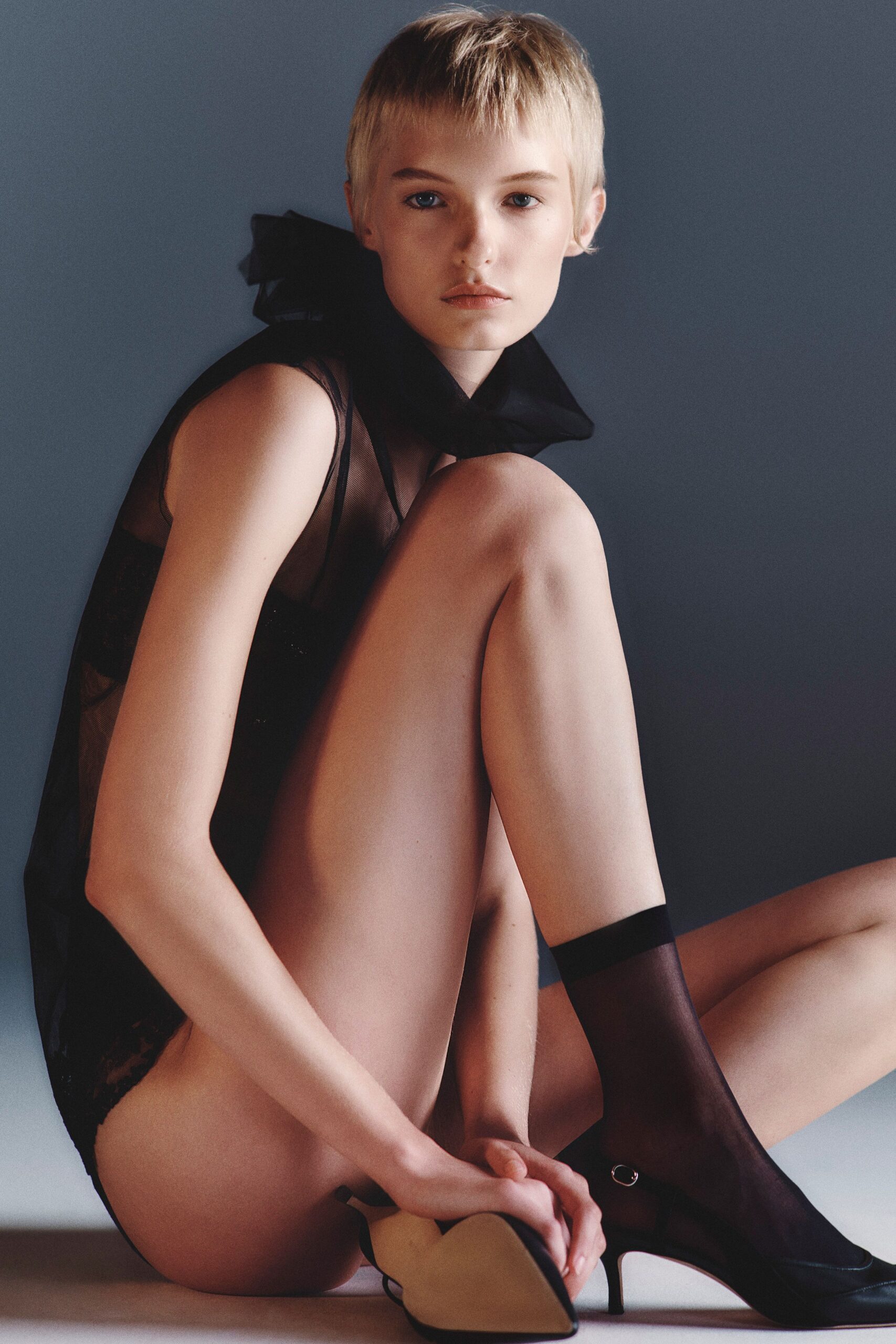 Sheer black mesh bodysuit over an opaque bandeau front. Oversized black bow standing stiff behind the neck. Smoke sheer ankle socks with a dense cuff band. Black pumps with a tiny silver ring at the vamp. Morning light on the hotel carpet after carrying your shoes upstairs.