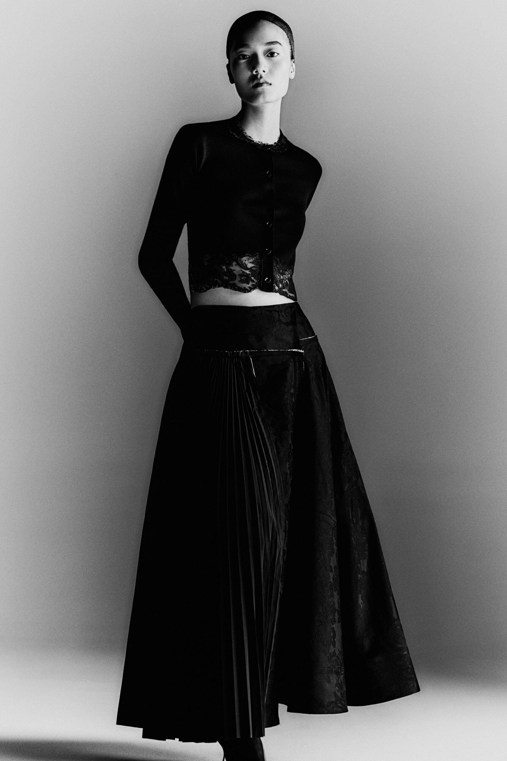 Black fine-knit cardigan with lace edging at neck and cropped hem. Asymmetric black skirt, one side knife-pleated, one side in shadowy floral jacquard. Pointed black boots just breaking the hem. After the string quartet, carrying your shoes across the museum courtyard.