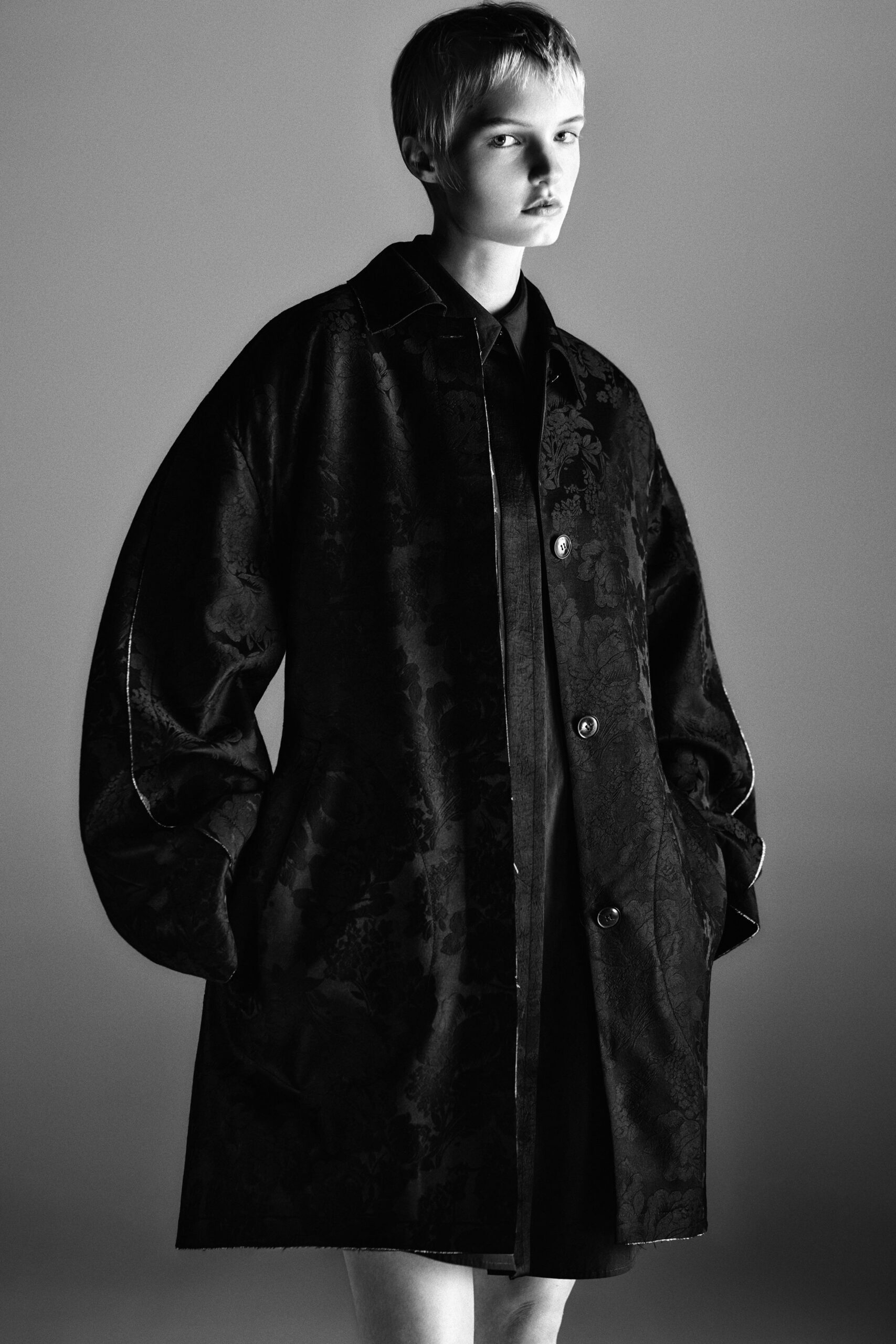 Black floral jacquard coat with balloon sleeves and a raw-cut hem. Black collared underlayer disappearing beneath the open placket. Museum atrium at closing, shoes echoing after the guard's last sweep.